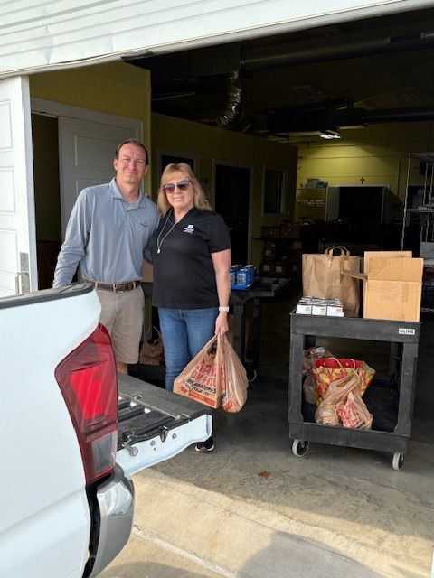 Two people stand in a garage next to a truck, one holding a grocery bag, with a utility cart of supplies nearby.