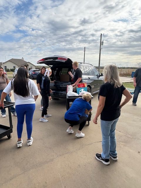 Volunteers unload boxes and bags from the back of a black SUV parked outdoors on a sunny day.