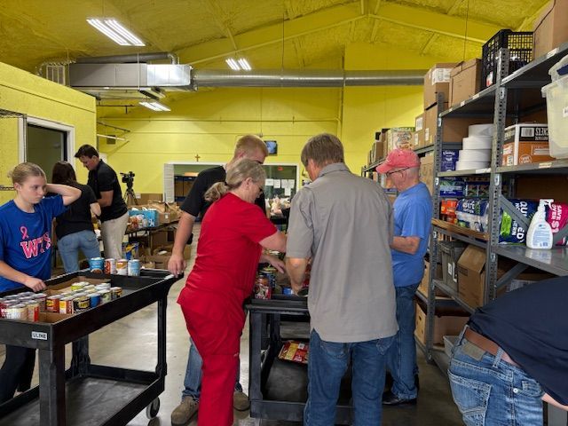 People sort and pack canned goods at a food pantry in a warehouse setting with yellow walls and metal storage shelves.