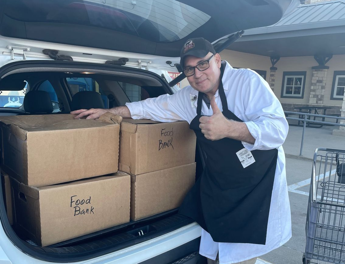 Person loading cardboard boxes into a car trunk at a parking lot, with a shopping cart nearby