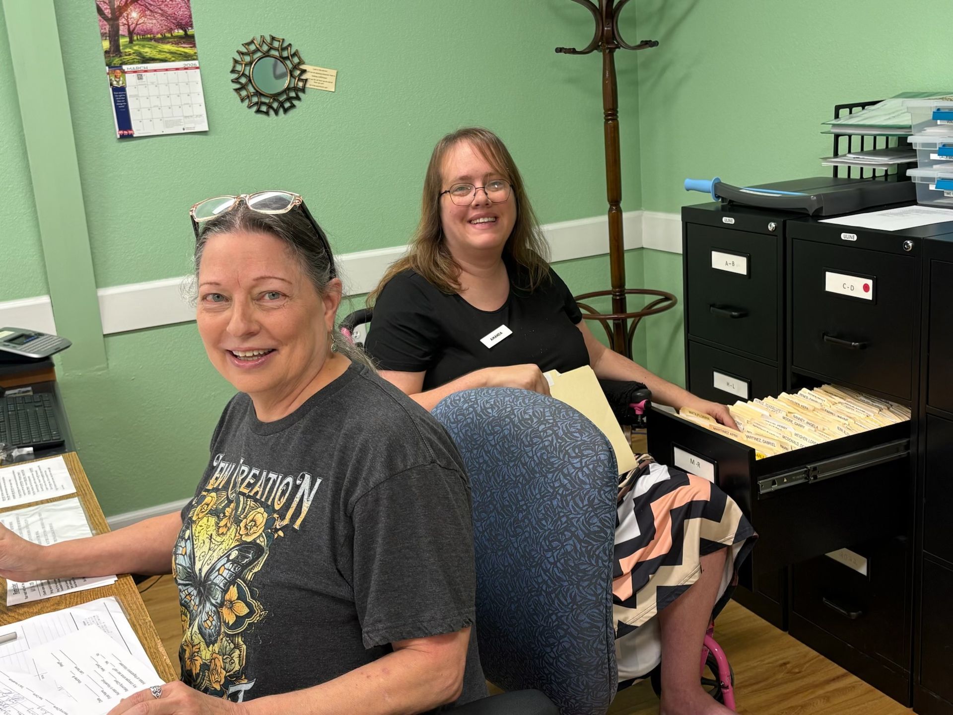 Two women sitting at desks in a green office, smiling at the camera, with filing cabinets and papers nearby.