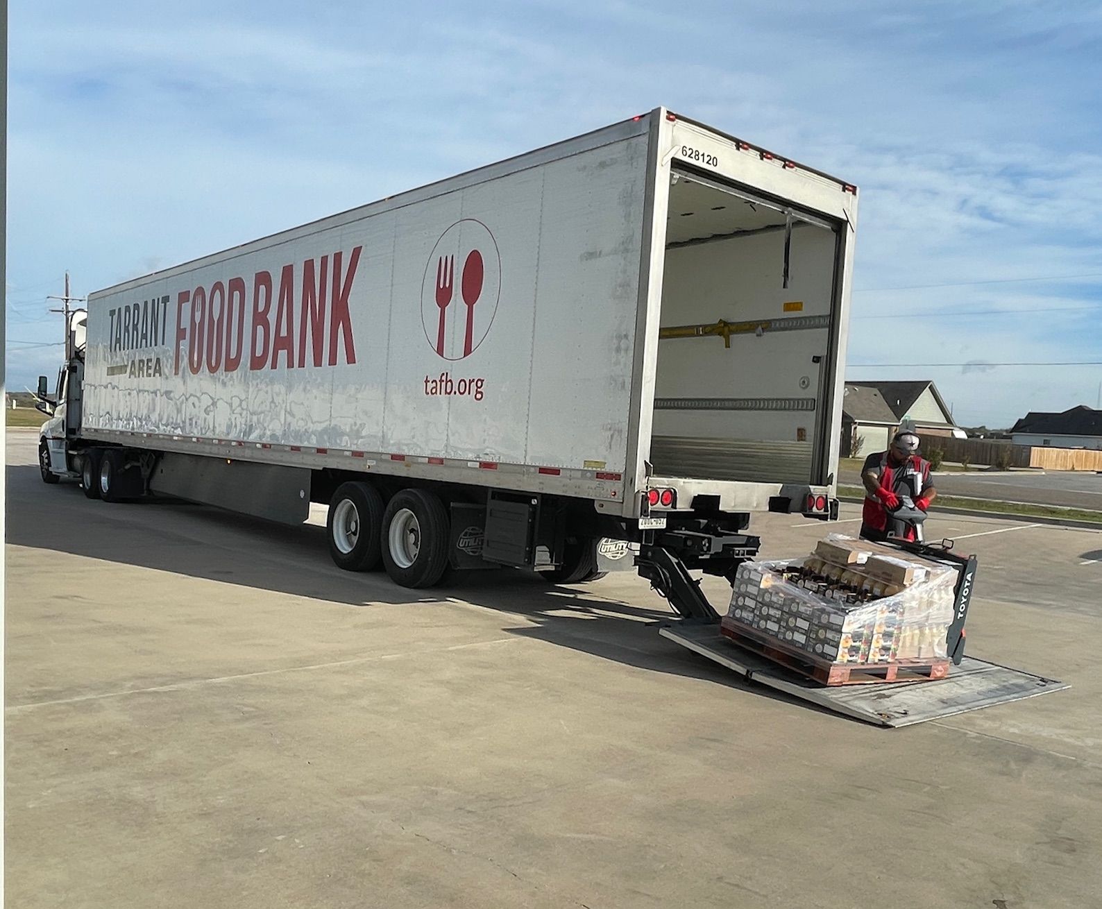 A food bank semi-truck parked outdoors as a worker uses a pallet jack to unload goods onto the liftgate.