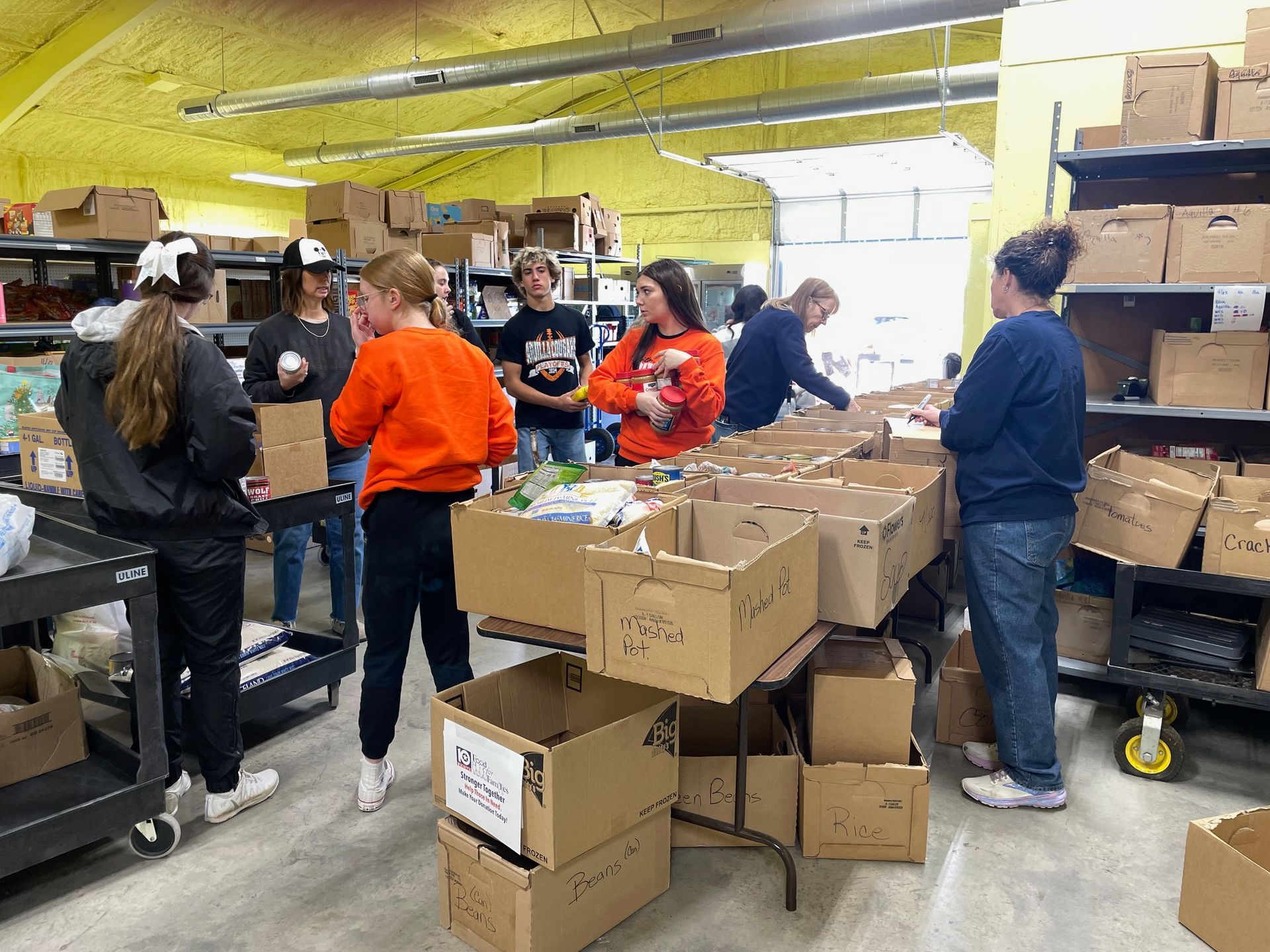 Volunteers sort items into cardboard boxes at a food pantry warehouse.