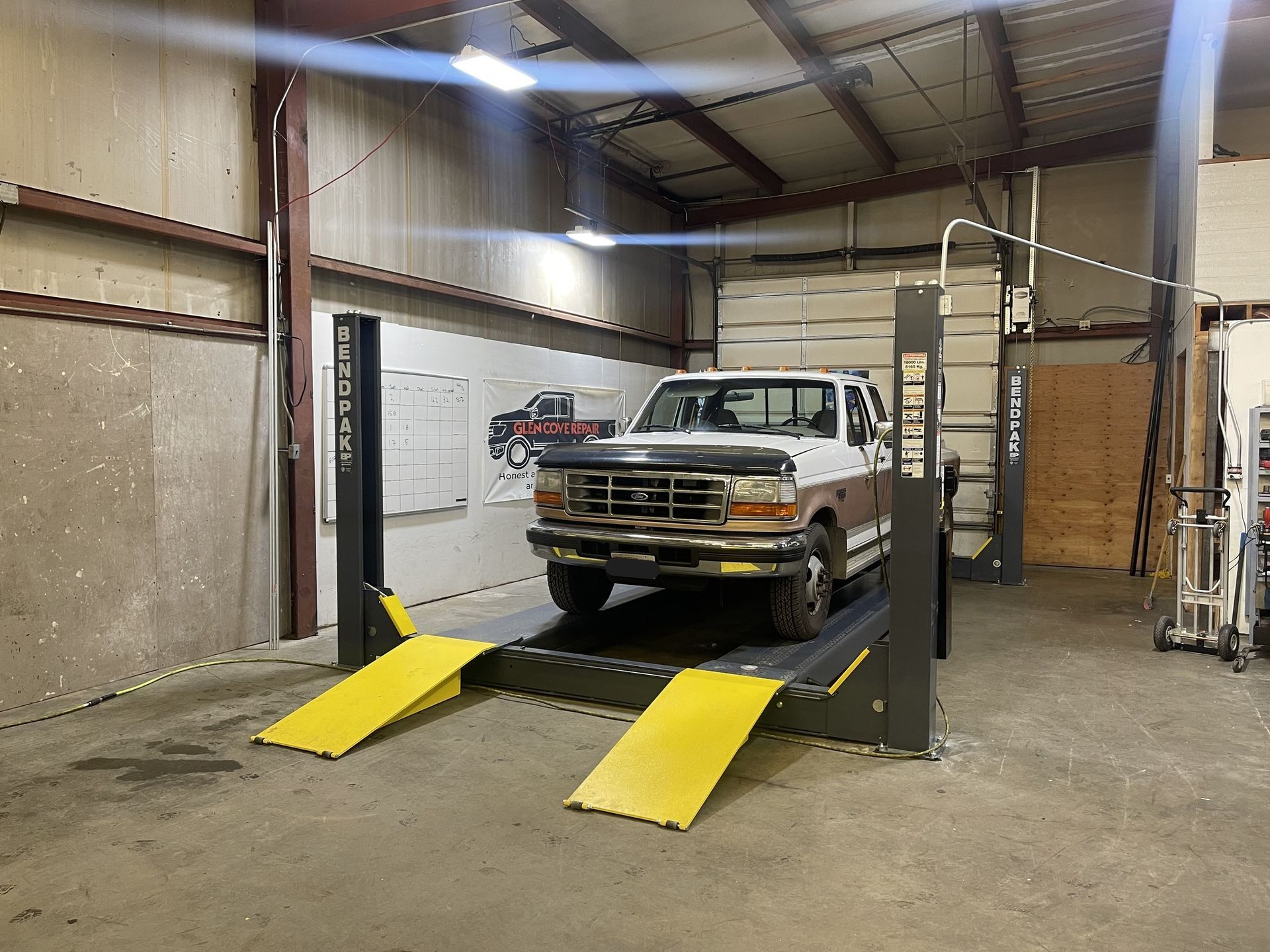 A white and brown pickup truck parked on a four-post vehicle lift inside an industrial repair garage. | Glen Cove Repair