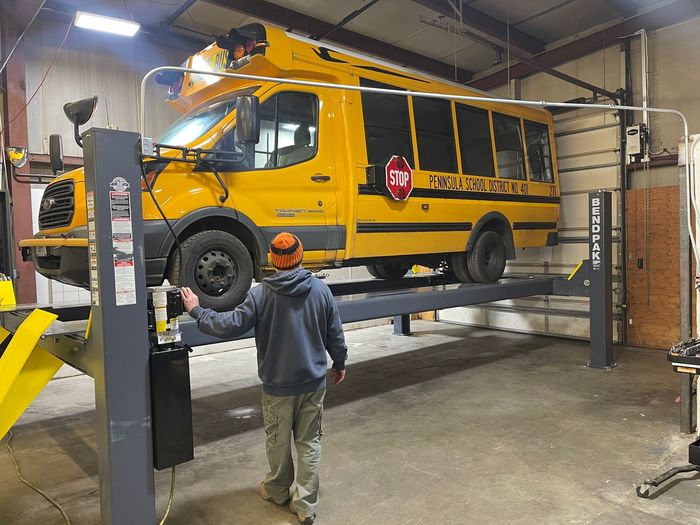 A person in a gray hoodie works in a garage on a yellow school bus lifted on a hydraulic hoist. | Glen Cove Repair