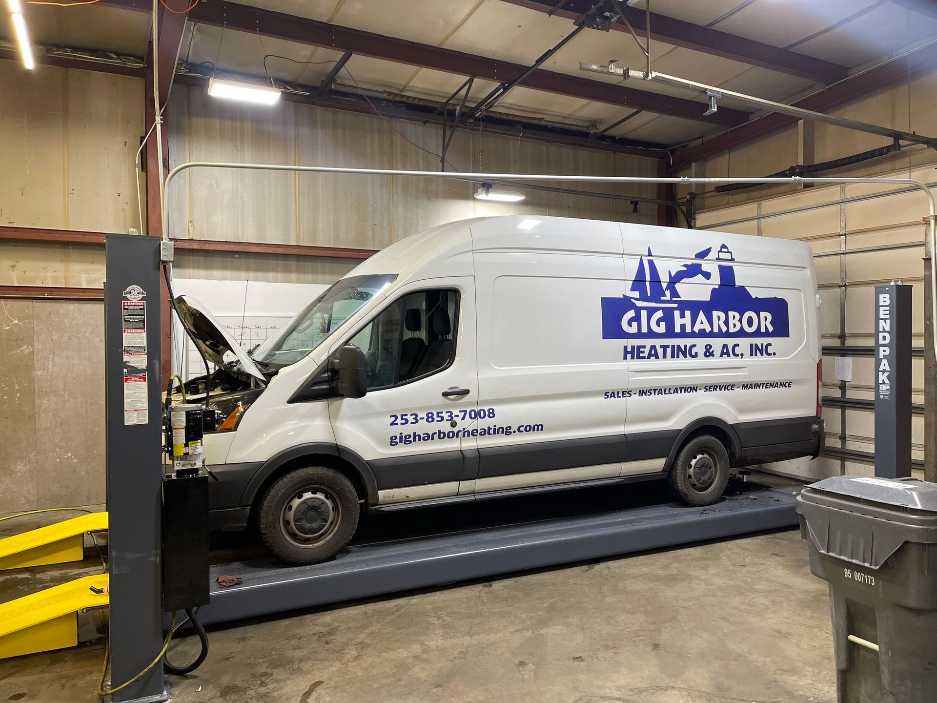 A white Gig Harbor Heating & A.C. service van with an open hood sits on an automotive lift inside a workshop. | Glen Cove Repair