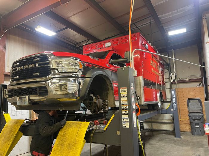 A mechanic works underneath a red ambulance raised on a lift inside a workshop. | Glen Cove Repair
