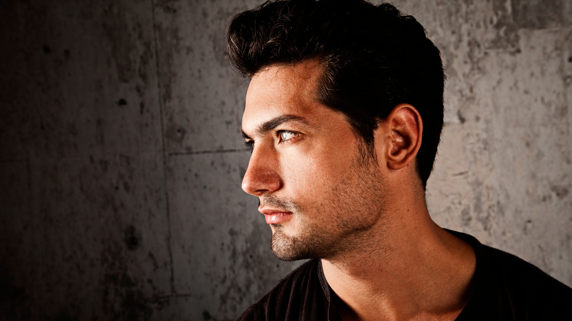 A man with a beard is standing in front of a wall and looking to the side.