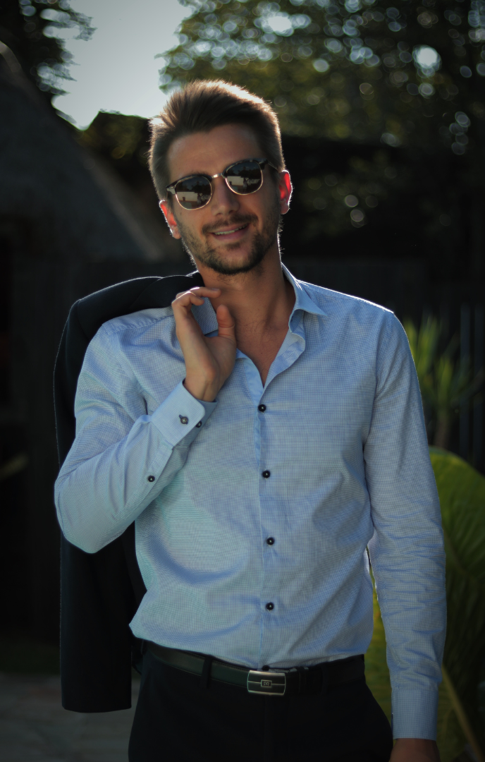 A man wearing sunglasses and a light blue shirt is holding his jacket over his shoulder