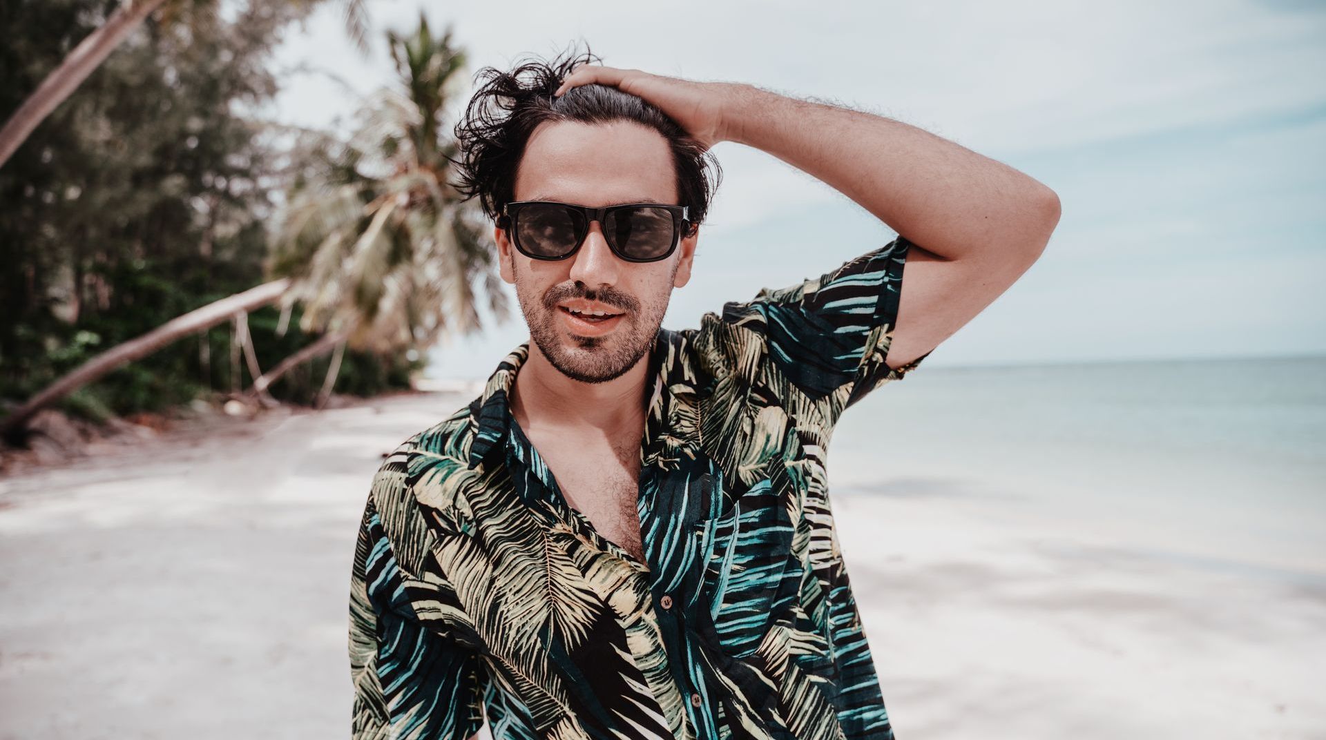 A man wearing sunglasses and a tropical shirt is standing on a beach.