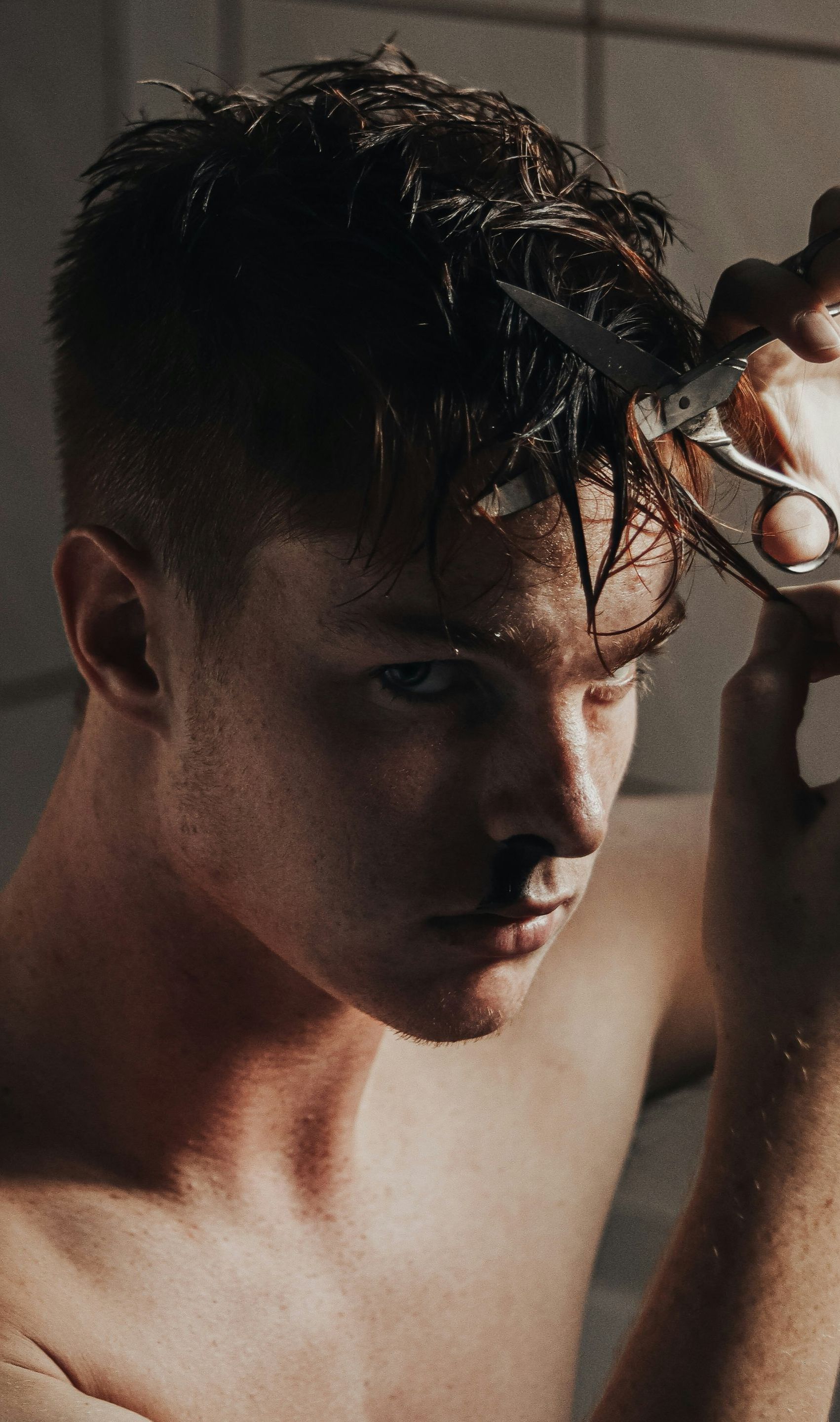 A shirtless man is cutting his hair with scissors in a bathroom.