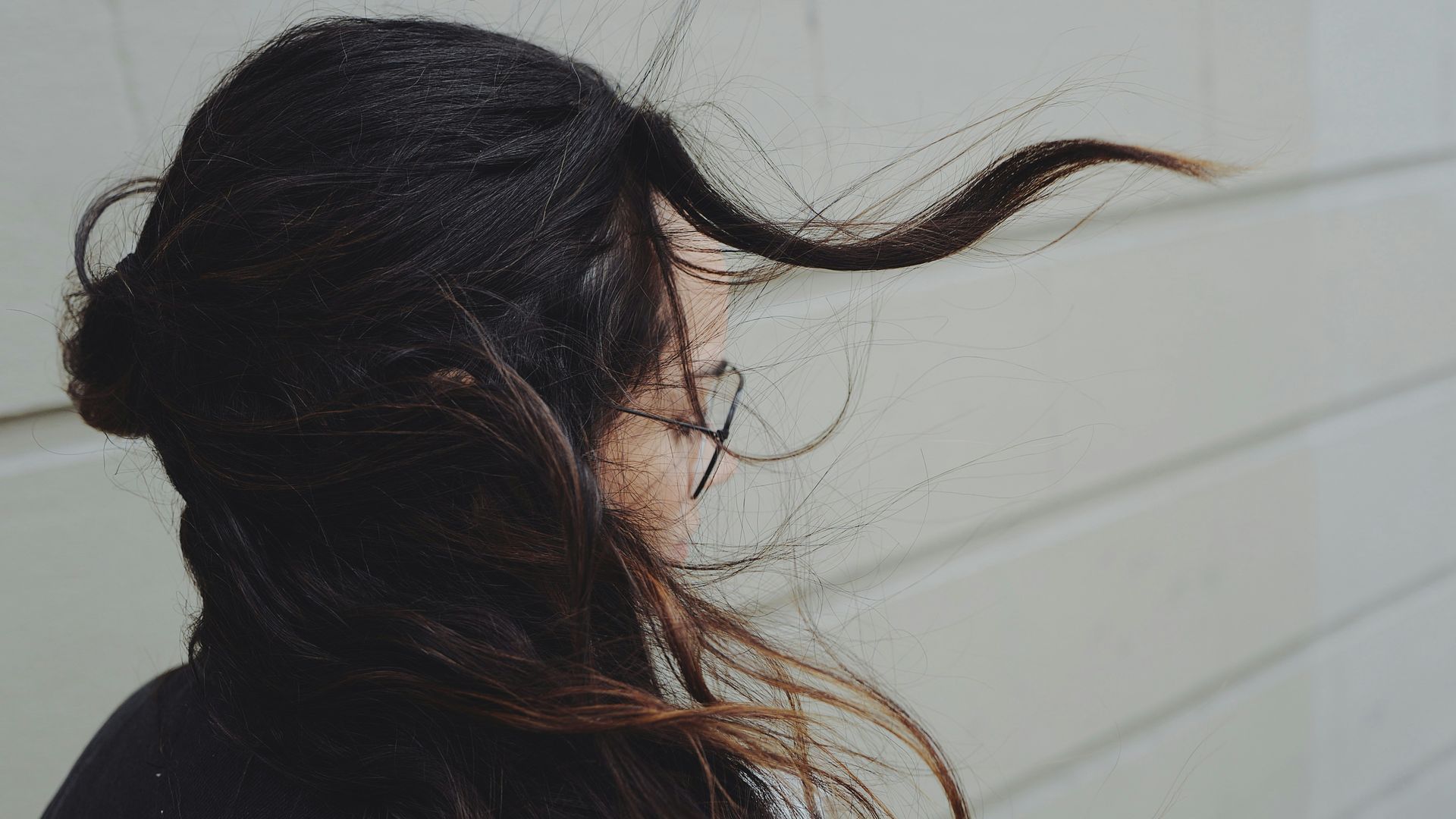 A woman with long hair is standing in front of a white wall with her hair blowing in the wind.