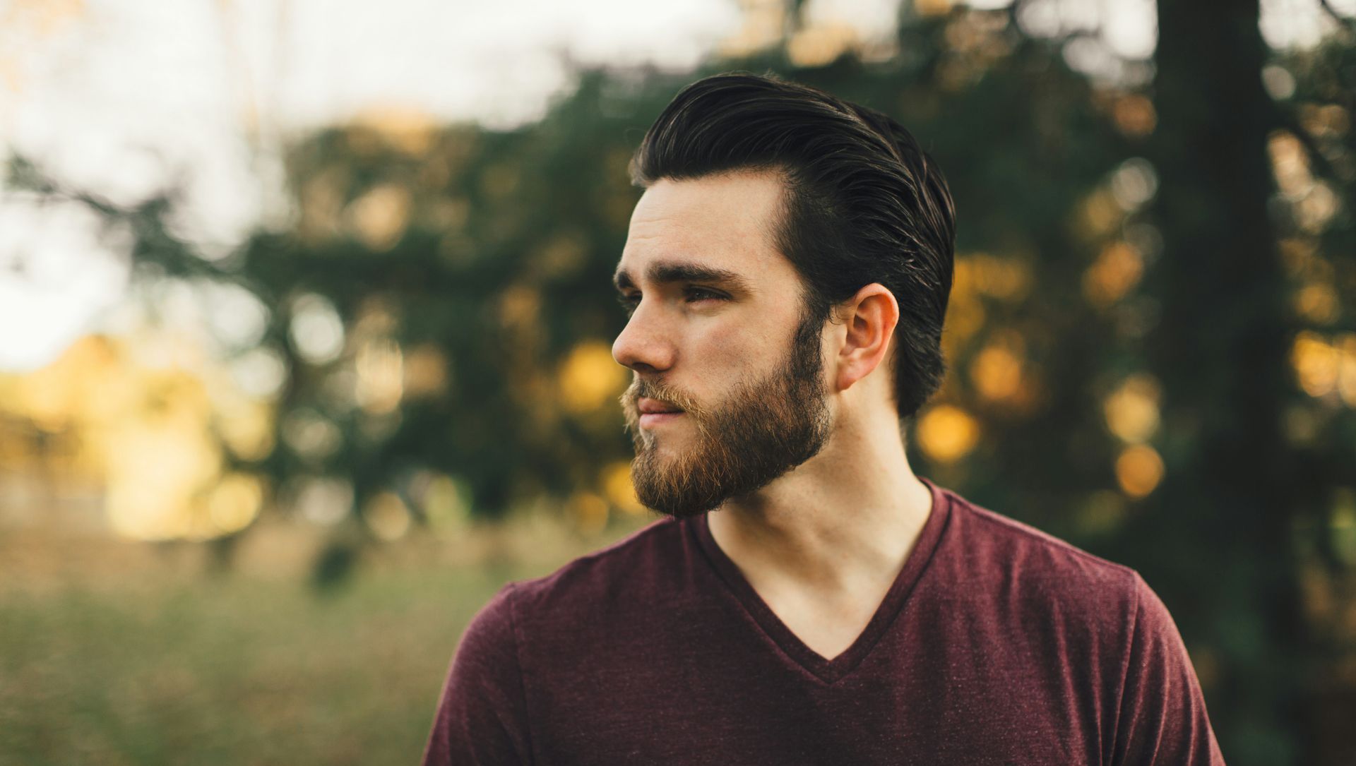A man with a beard is wearing a maroon shirt and standing in the woods.