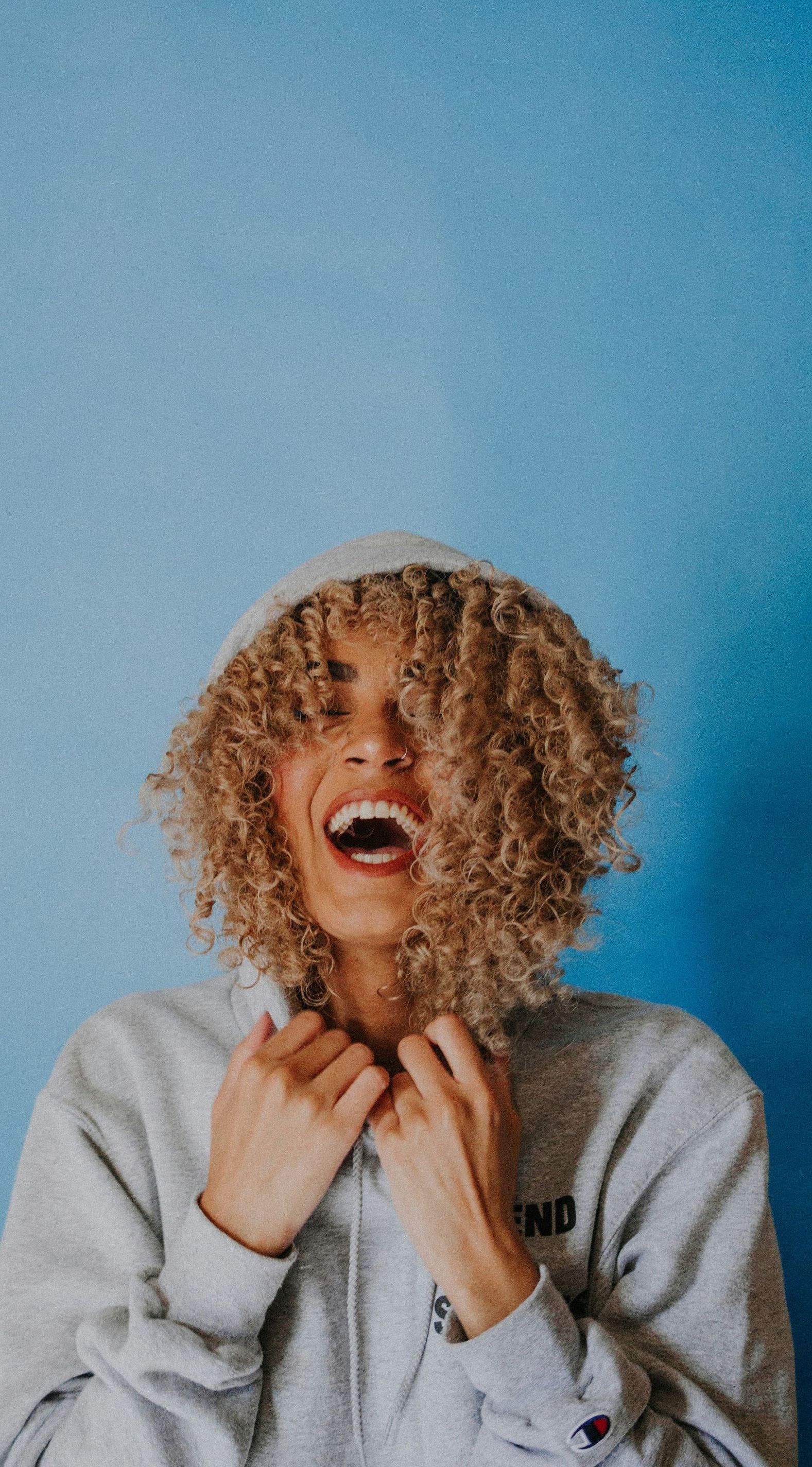 A woman with curly hair is wearing a hoodie and laughing.