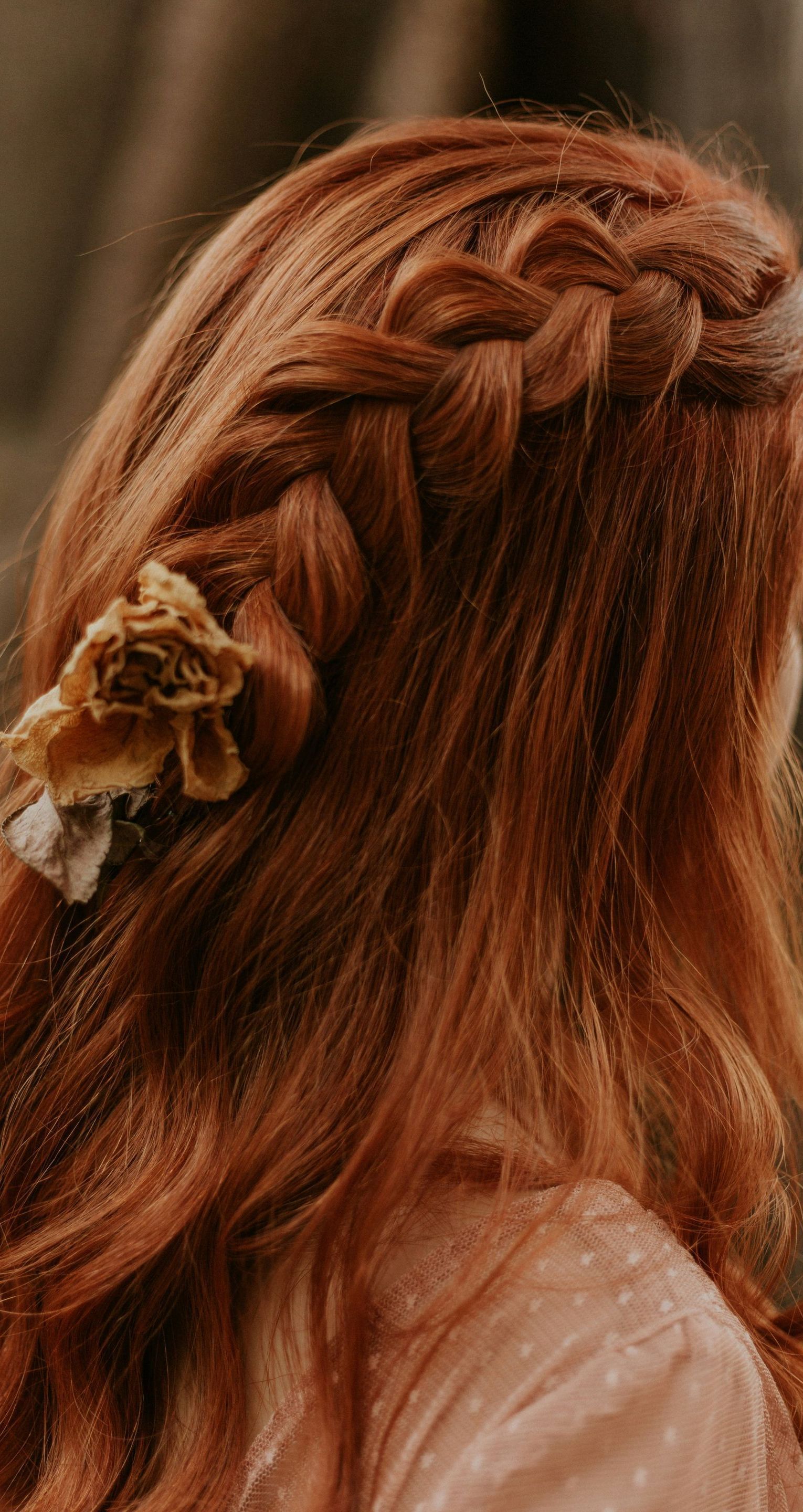 A woman with red hair is wearing a braided hairstyle with a flower in her hair.