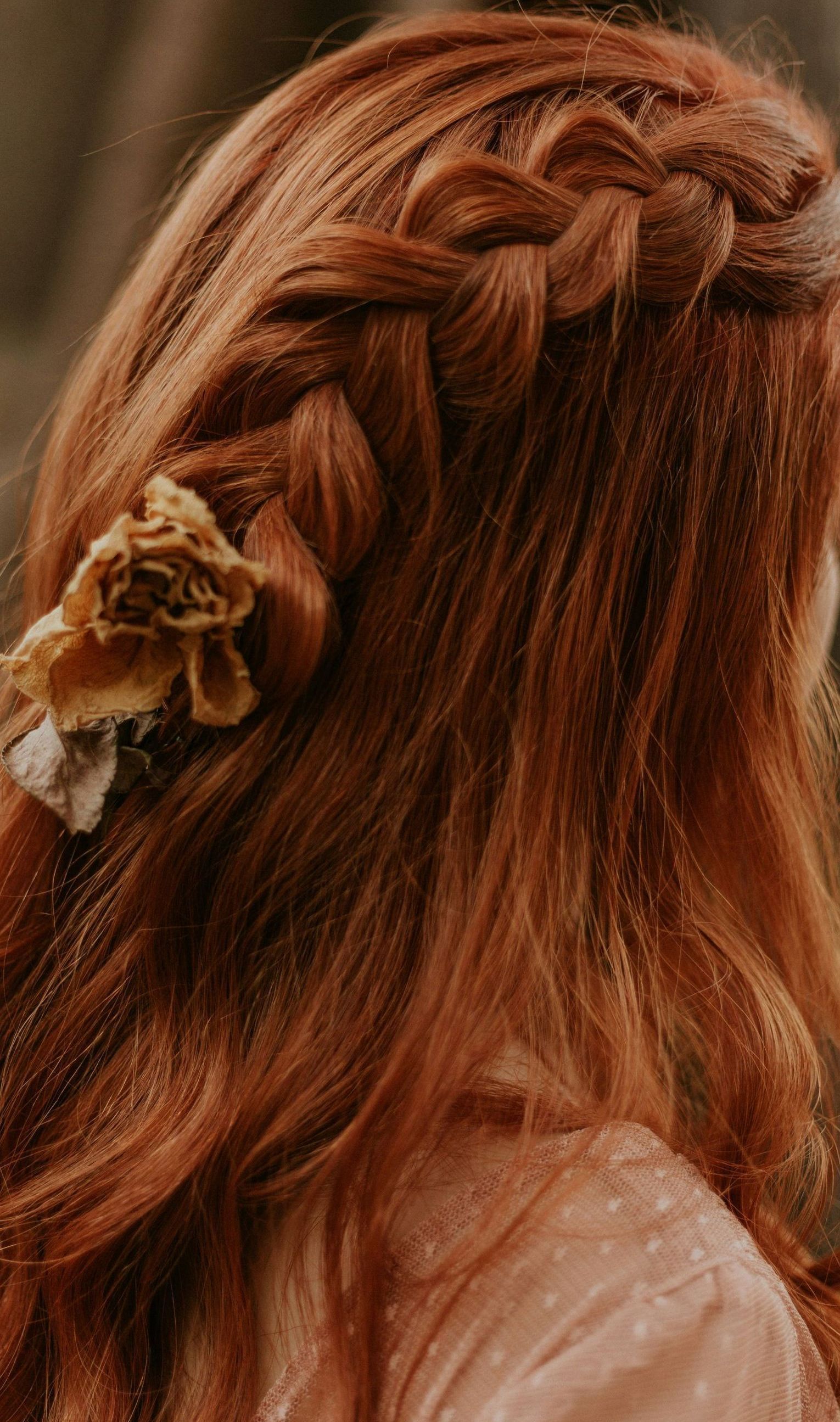 A woman with red hair is wearing a braided hairstyle with a flower in her hair.