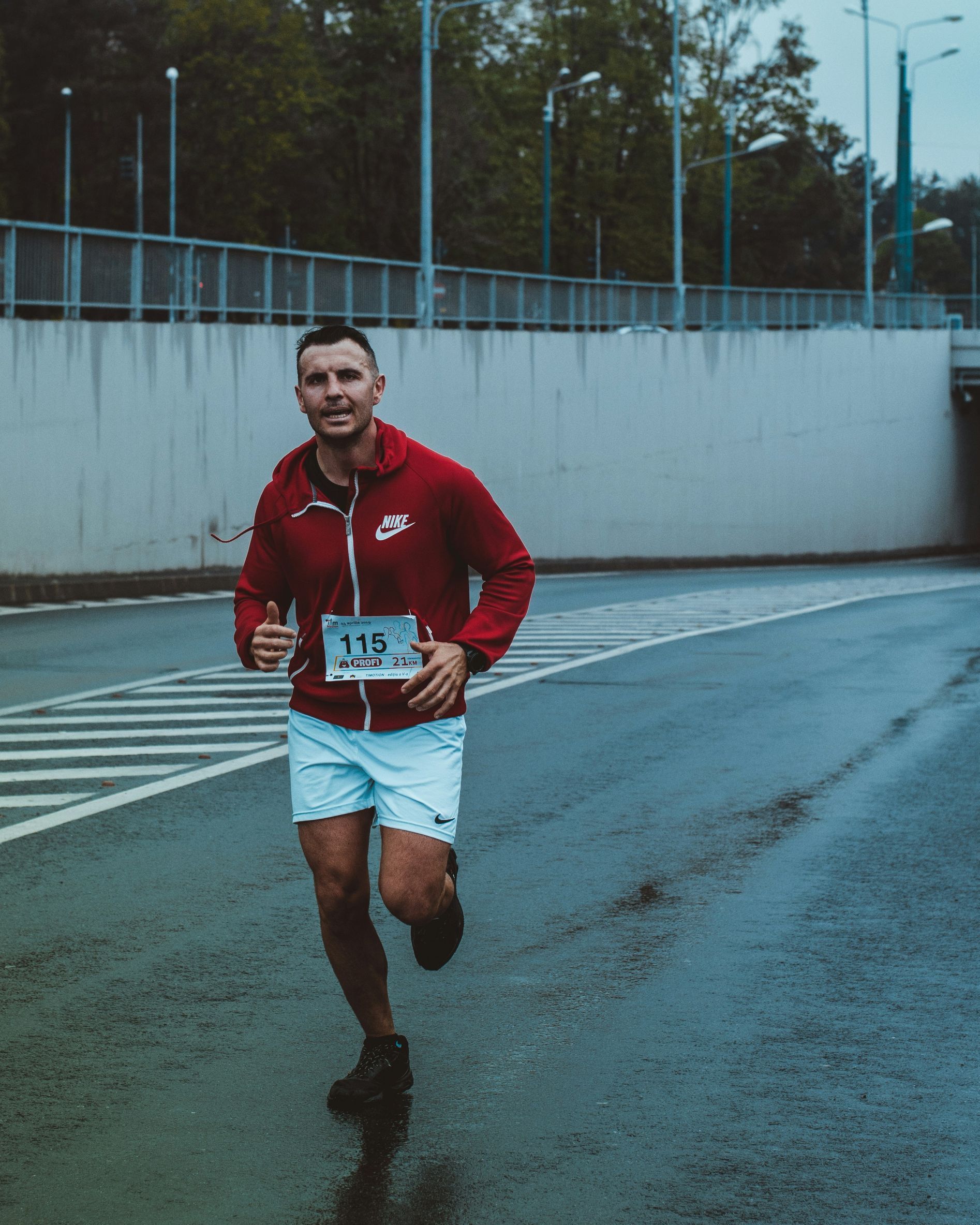 A man in a red jacket and white shorts is running on a wet road