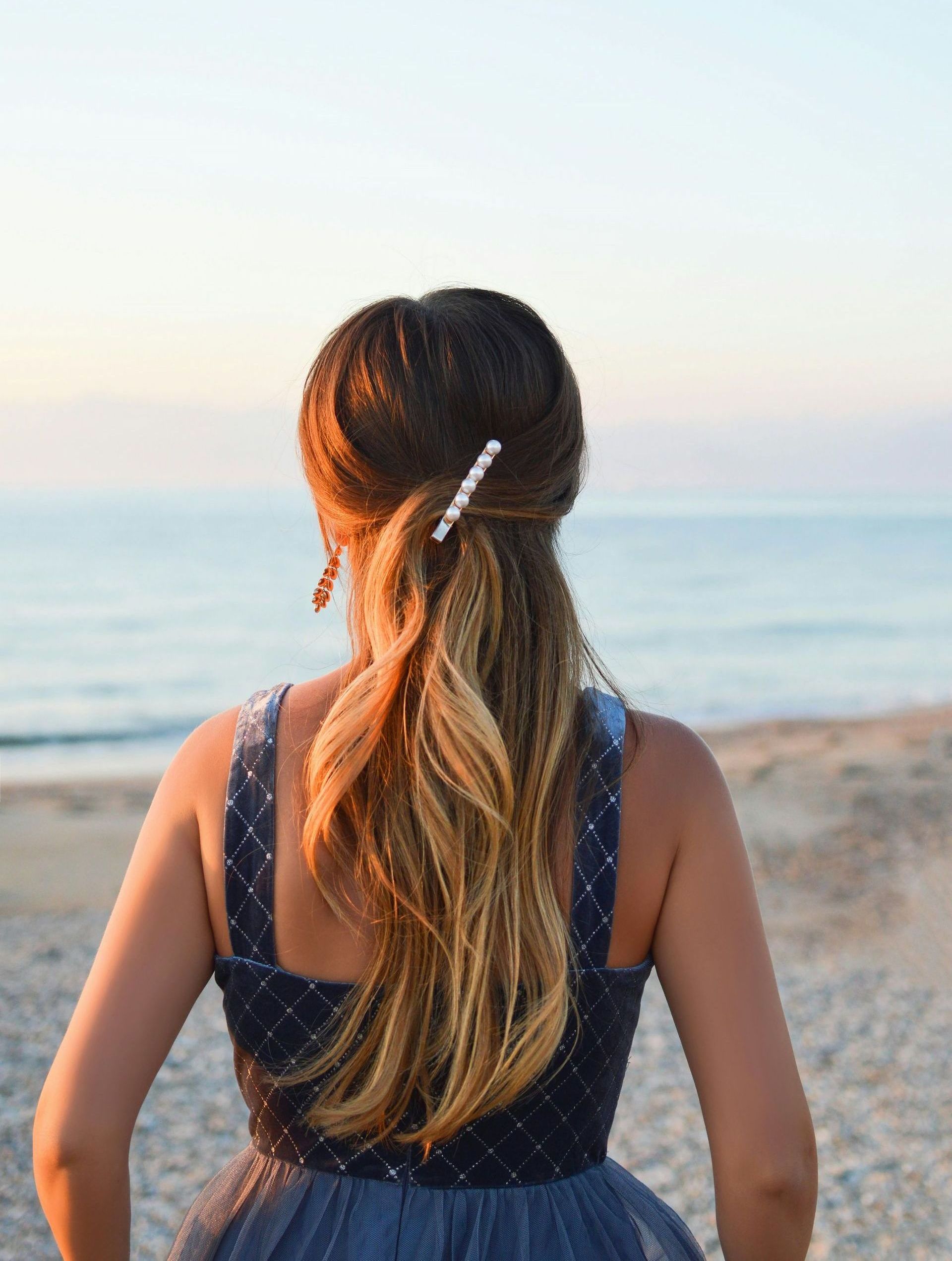 A woman in a blue dress is standing on the beach looking at the ocean.