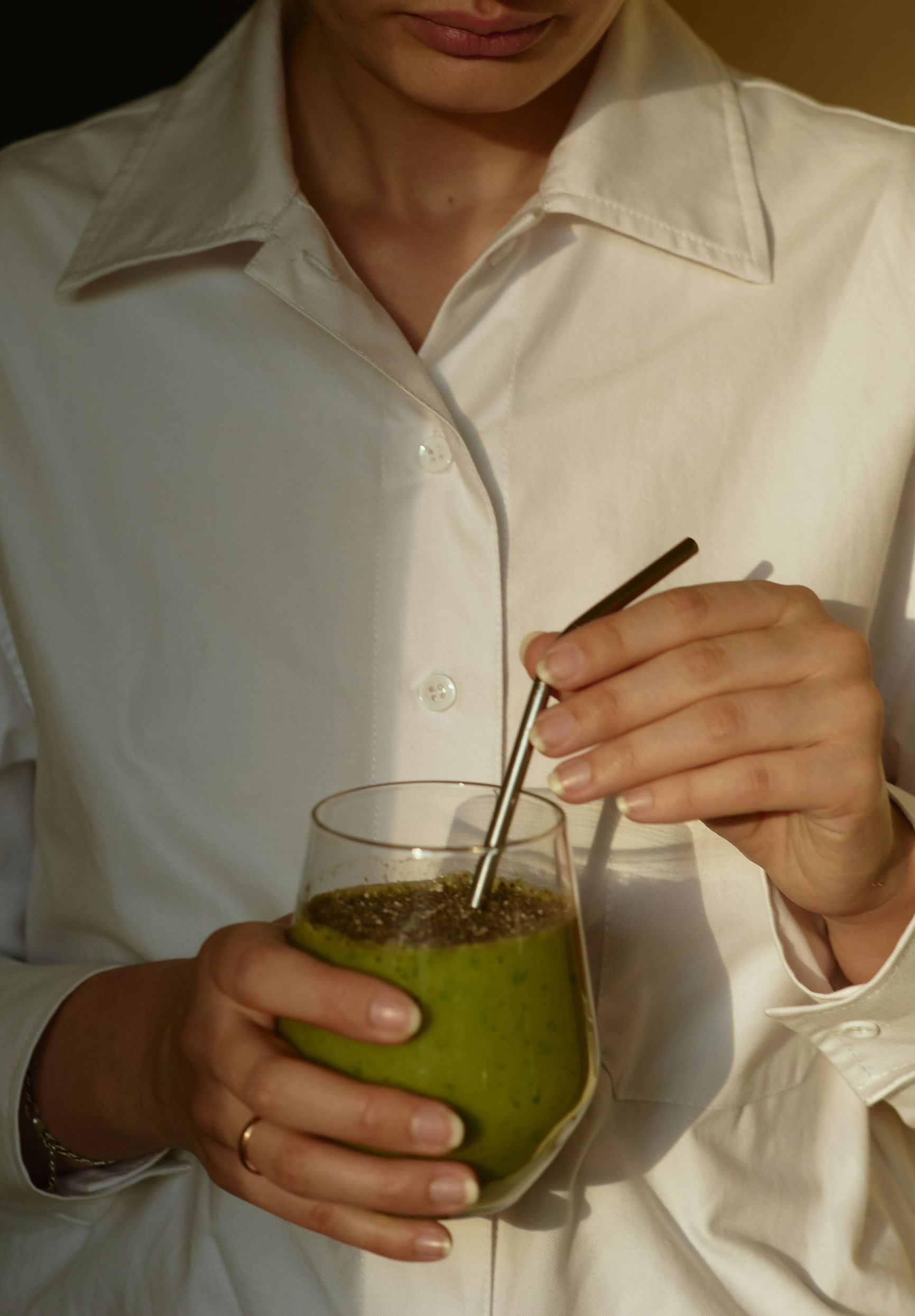 Person in a white shirt holds a glass of green smoothie with a metal straw.