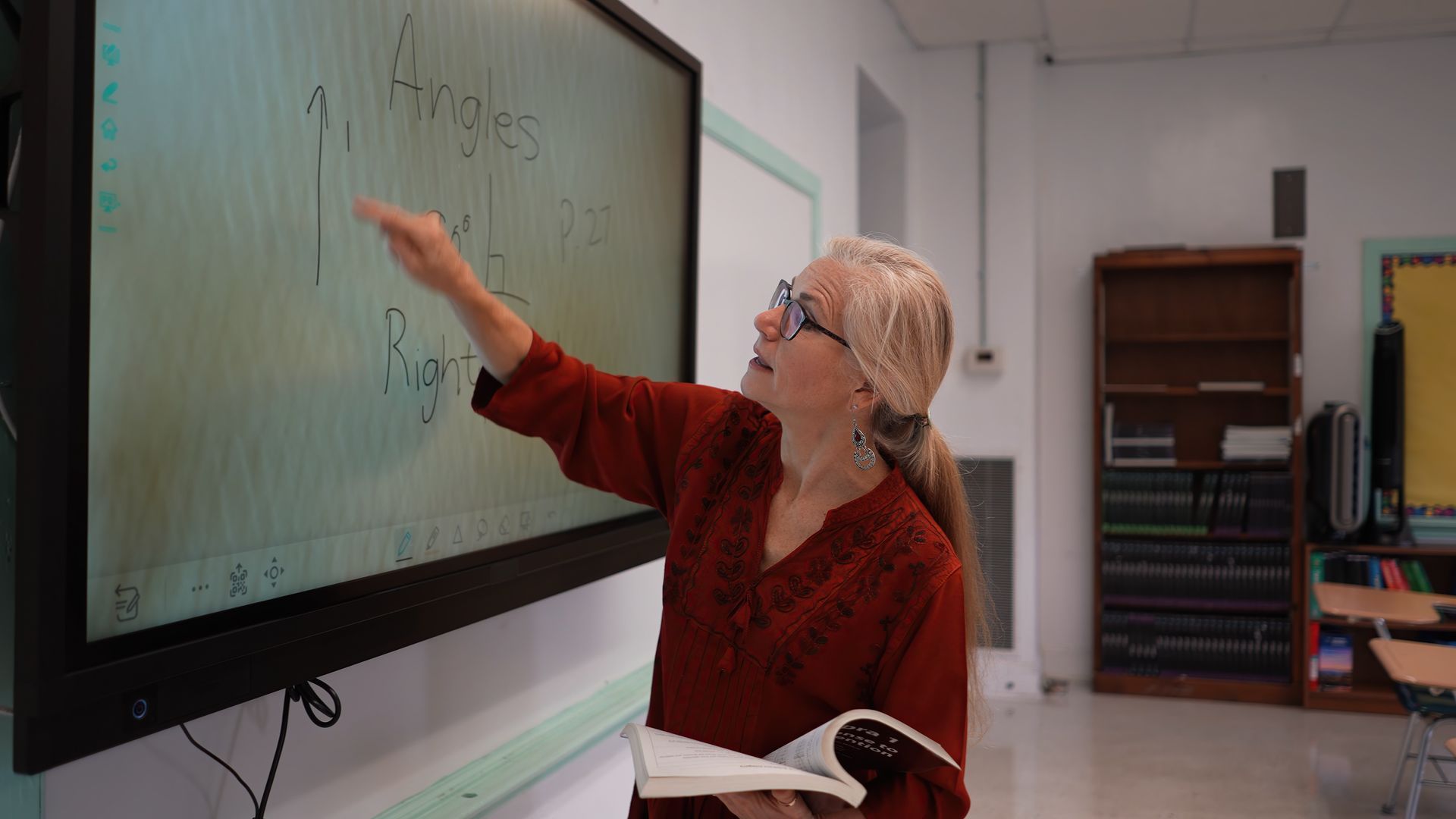 Person pointing at a digital board displaying notes about angles while holding an open book.
