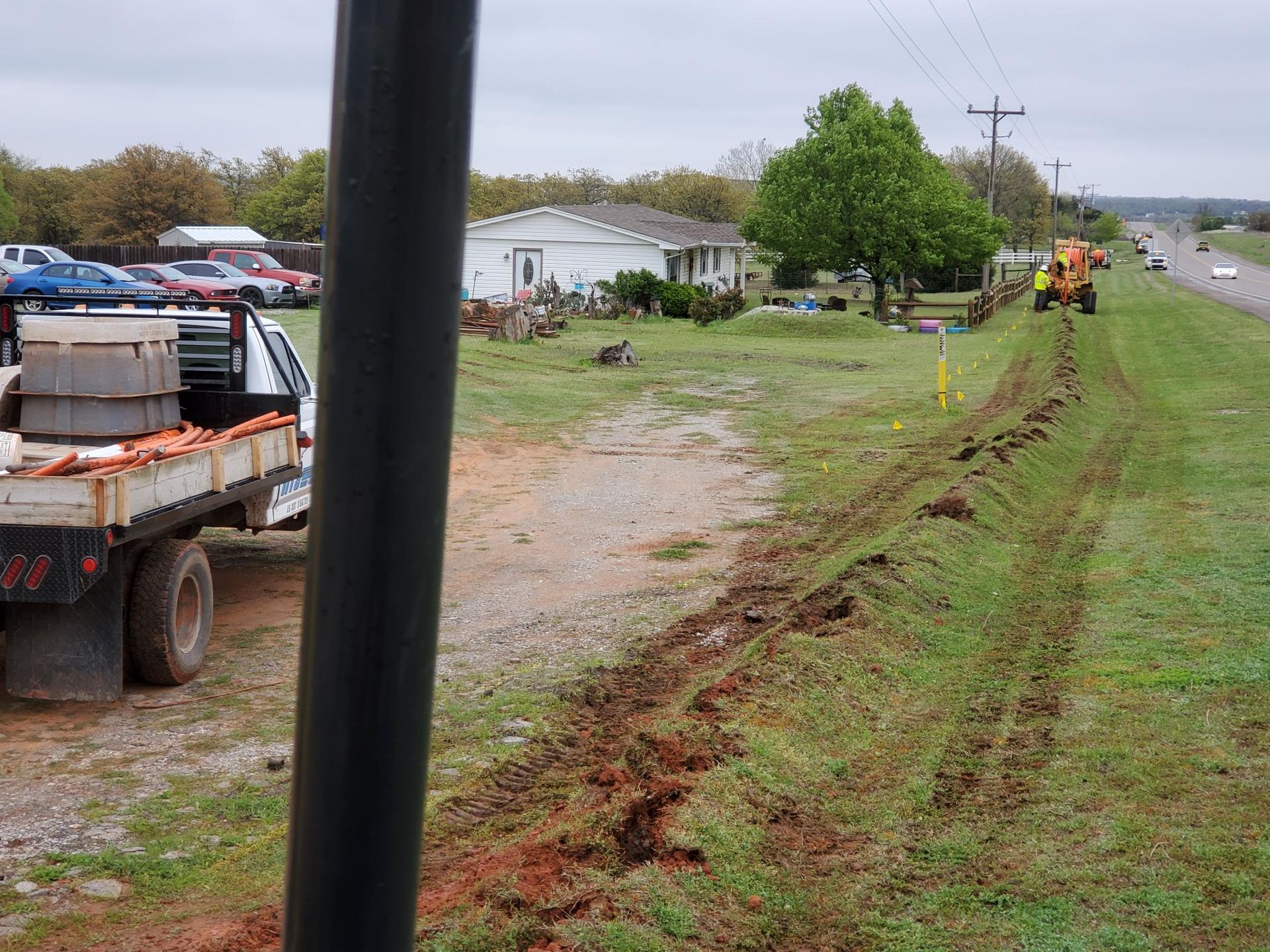 A trench being dug in grass next to a road, with a truck and house in the background.