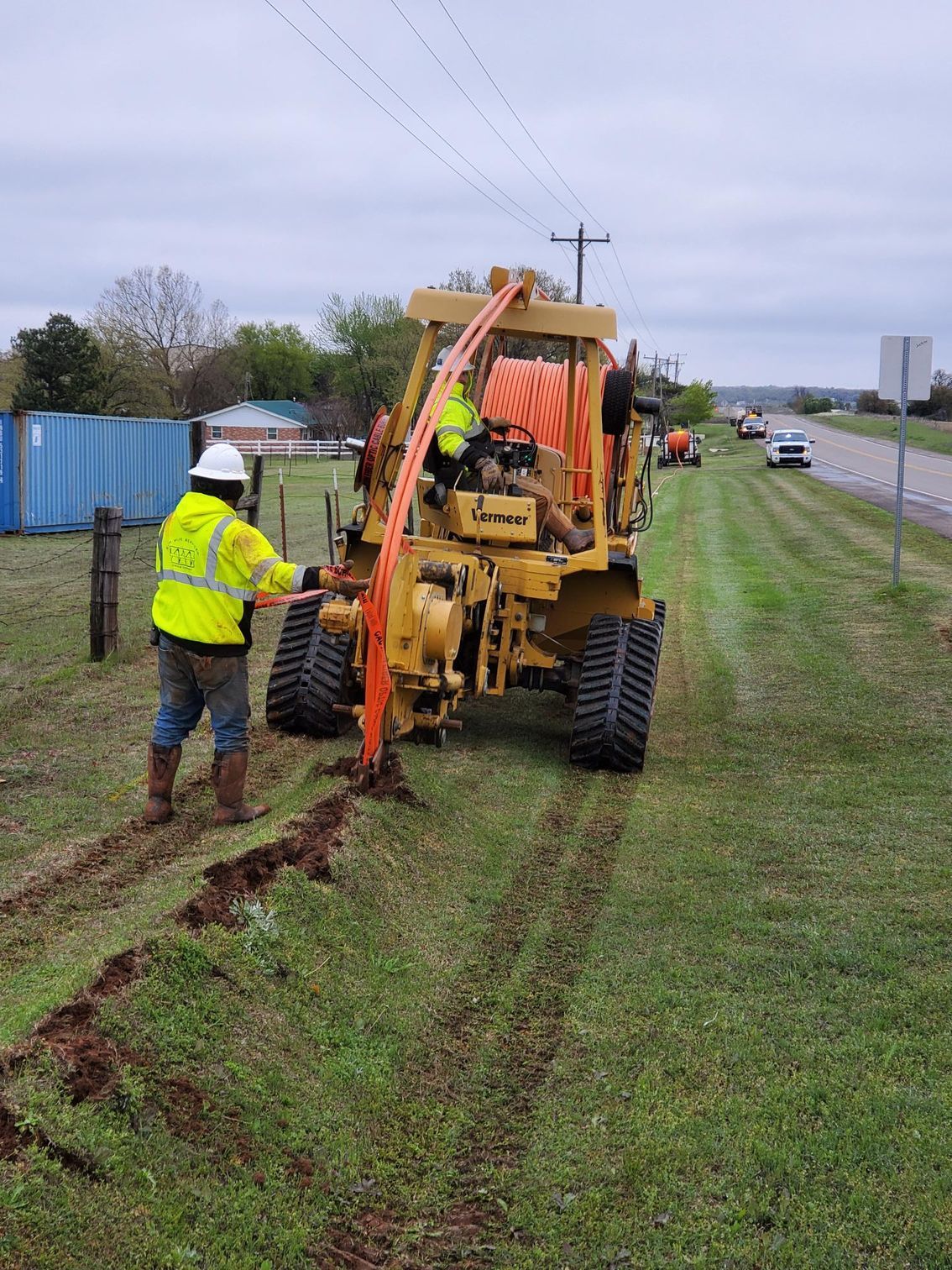 Workers installing underground utilities using a trenching machine next to a road.