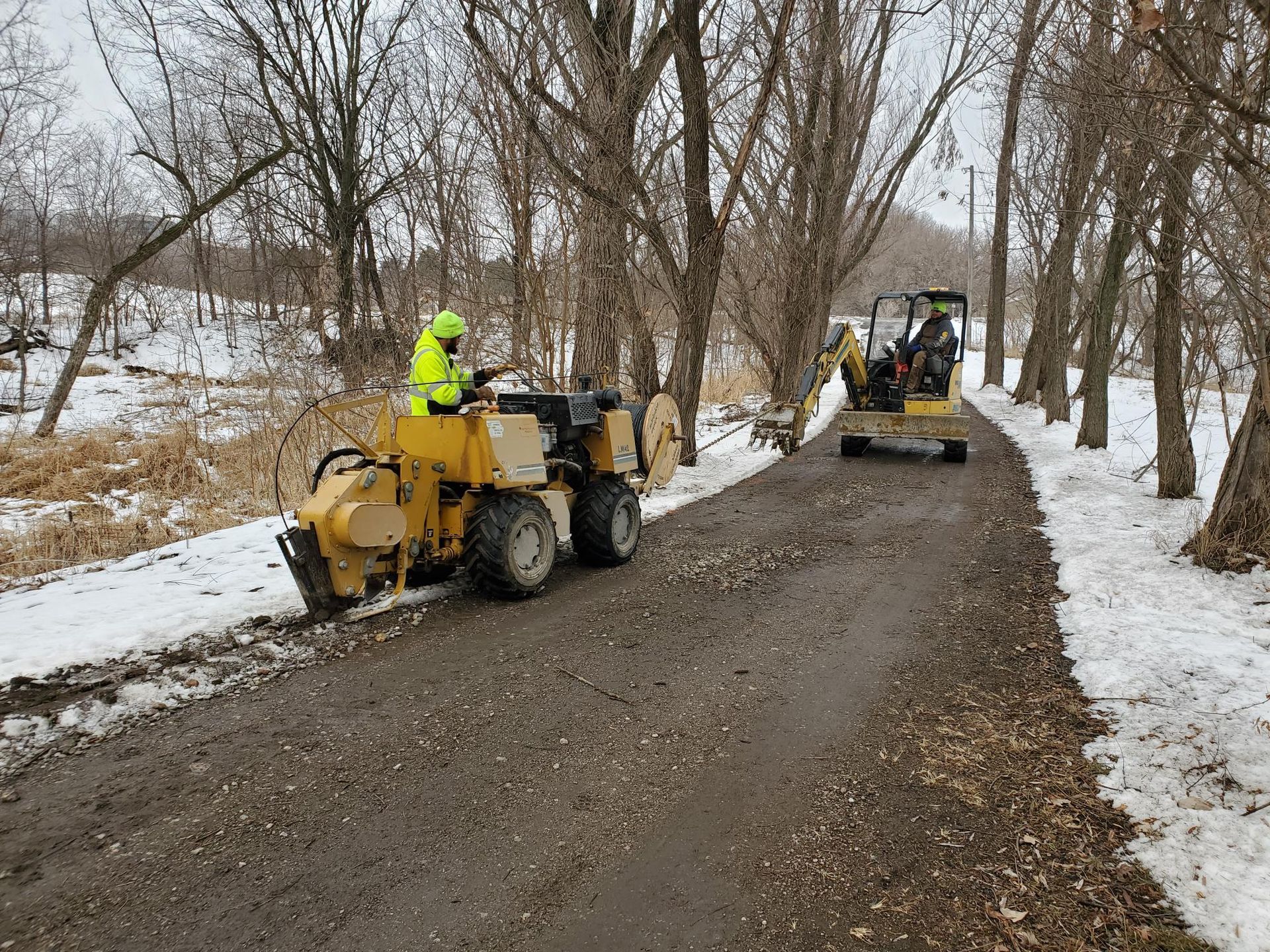 Two machines working on a path next to trees, snowy ground. One operator wears a reflective vest.