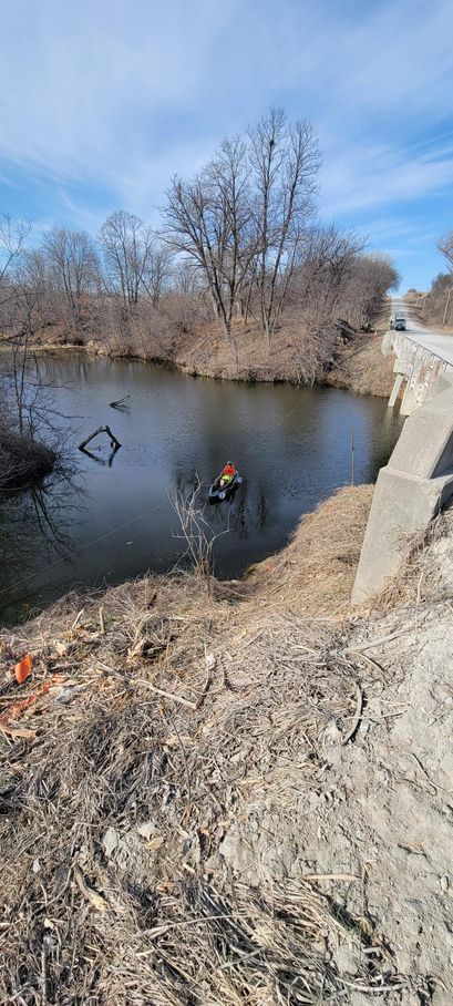 A person in a boat on a river, under a bridge on a sunny day.