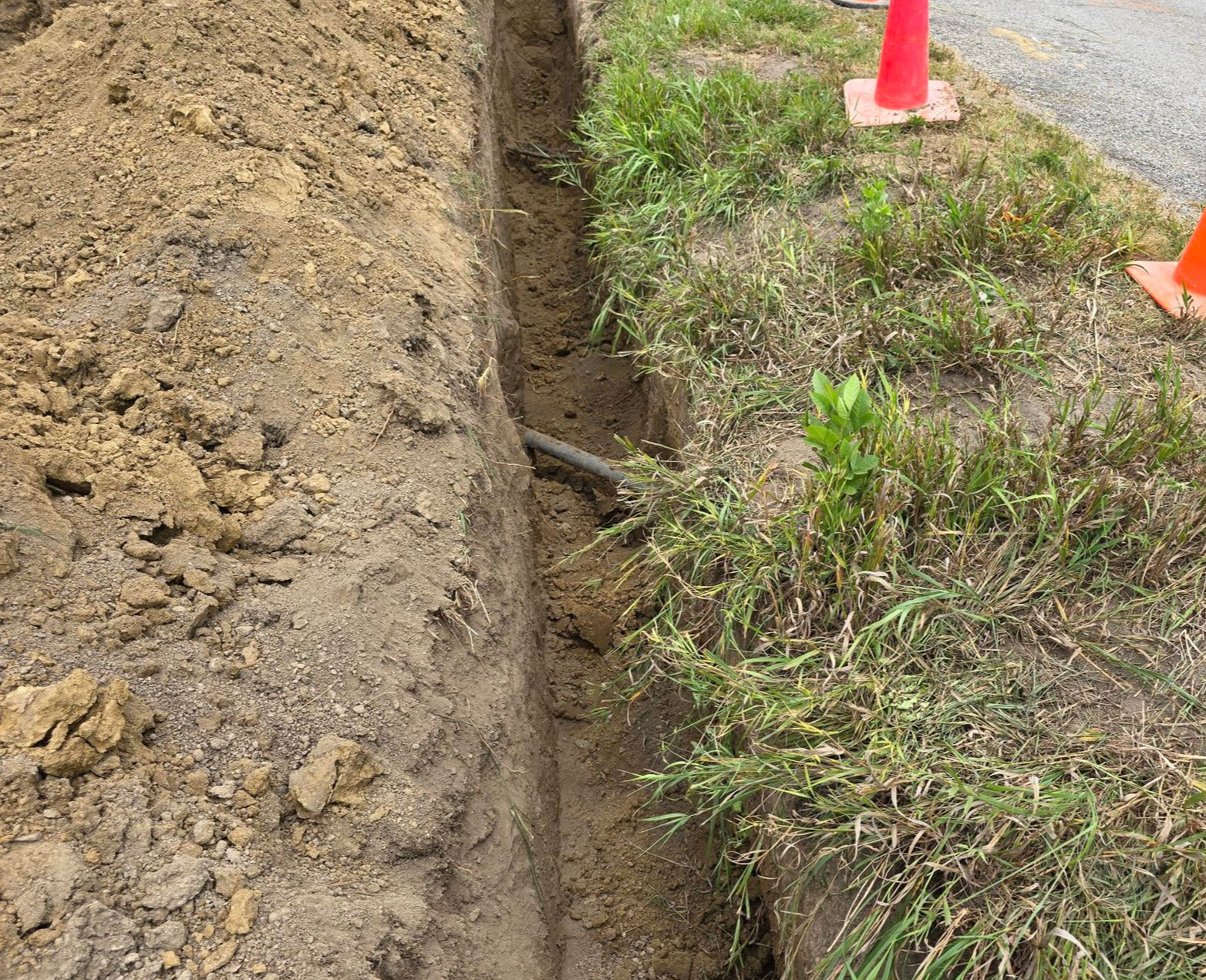 Narrow trench dug in dirt beside grass and road, with orange traffic cones.