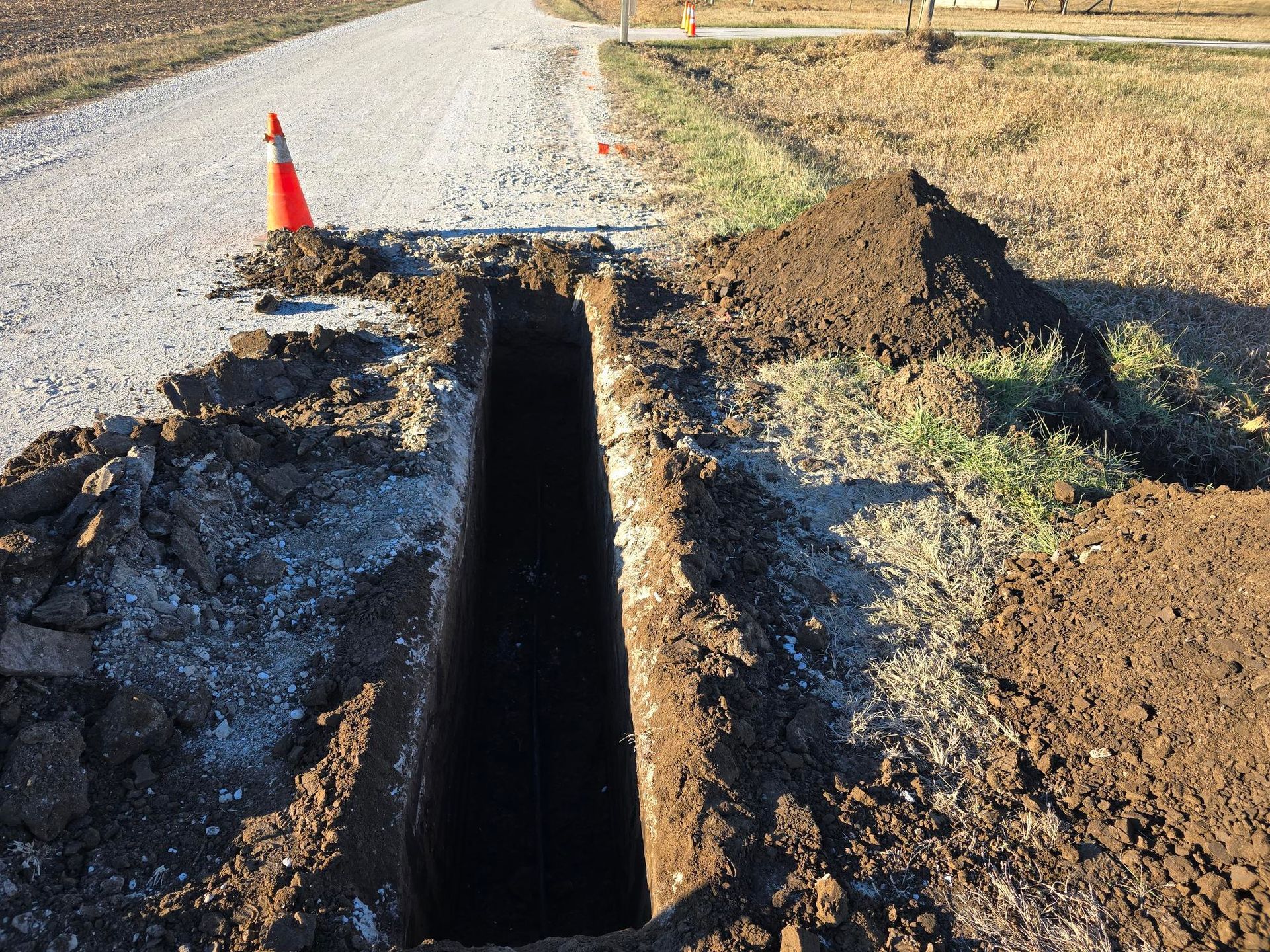 A deep trench dug across a gravel road, with an orange traffic cone visible.