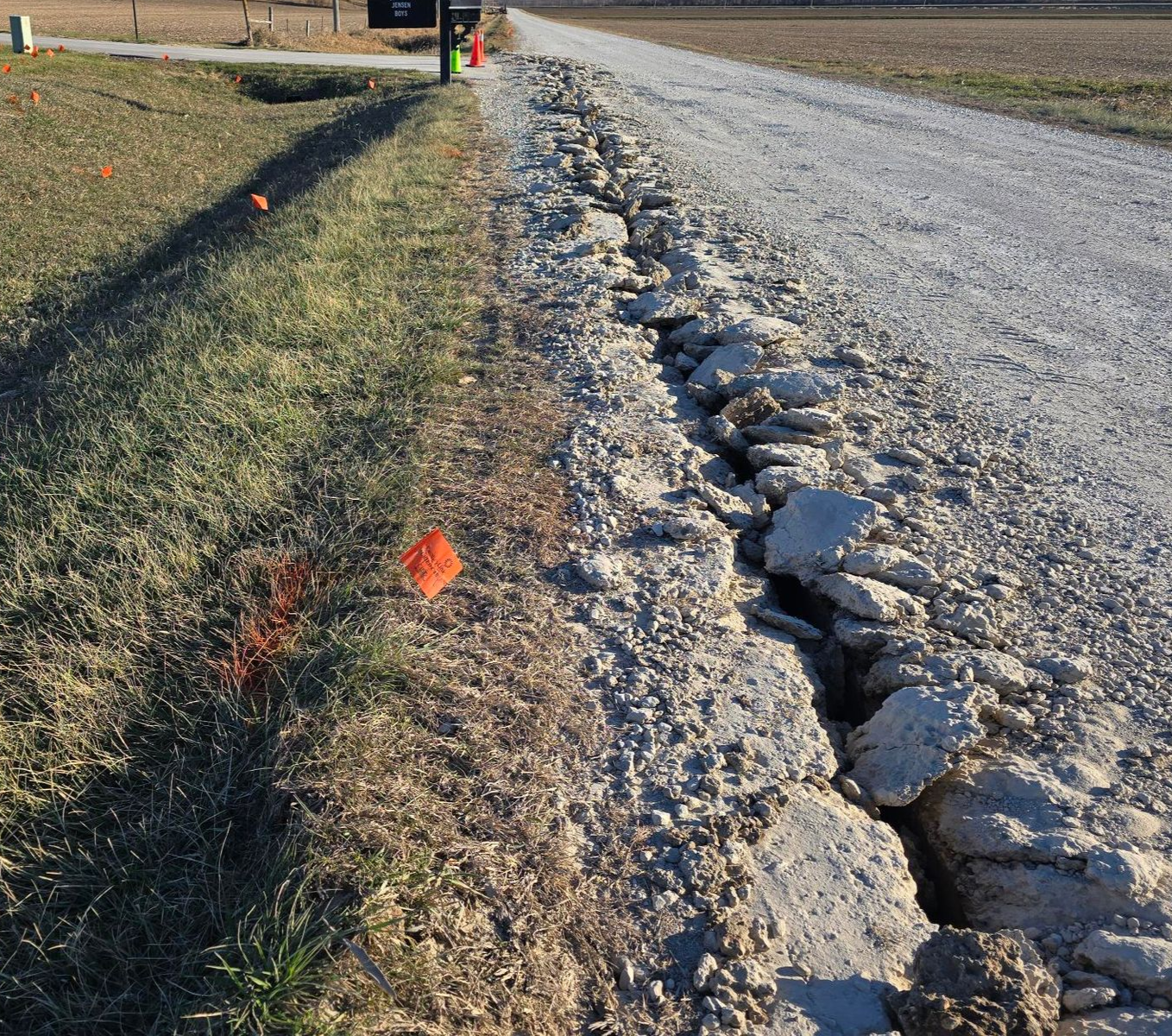 Gravel road with a large crack running along its side, next to grassy verge. Orange markers are visible.