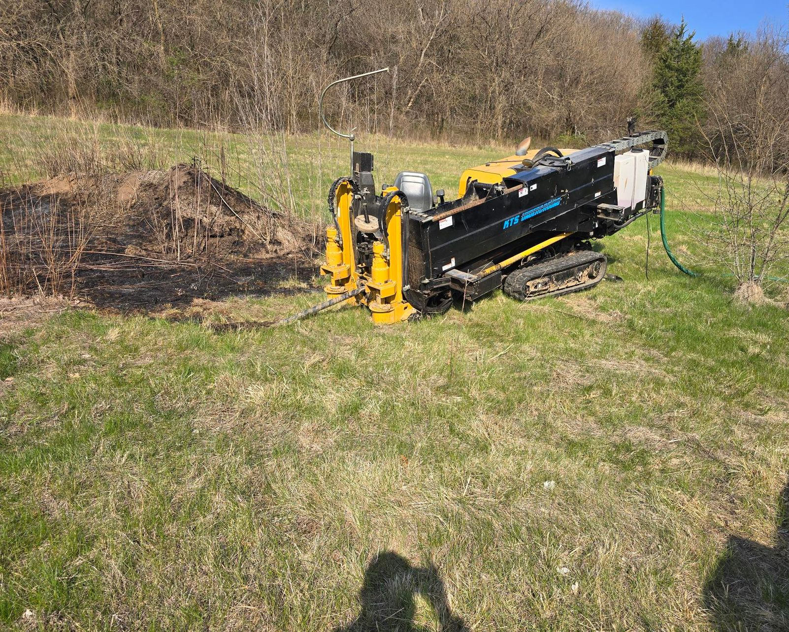A small yellow and black drilling machine on a grassy hill, with a treeline in the background.