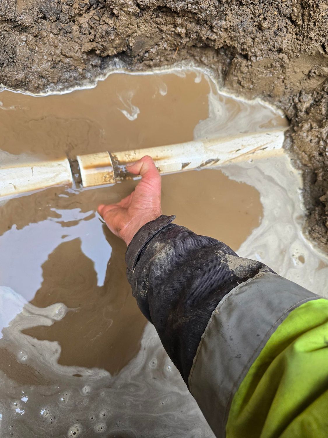 Hand touching a broken white pipe in muddy water within a trench.