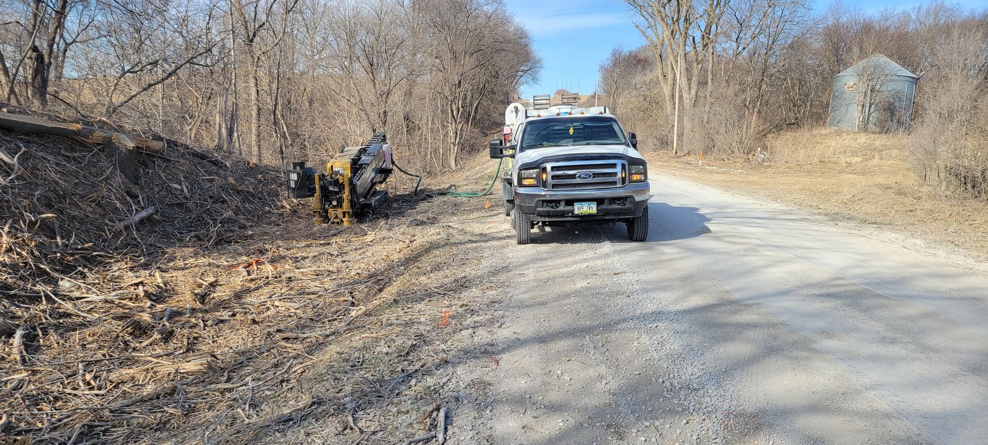 A truck and equipment working on a gravel road, with trees on the sides.