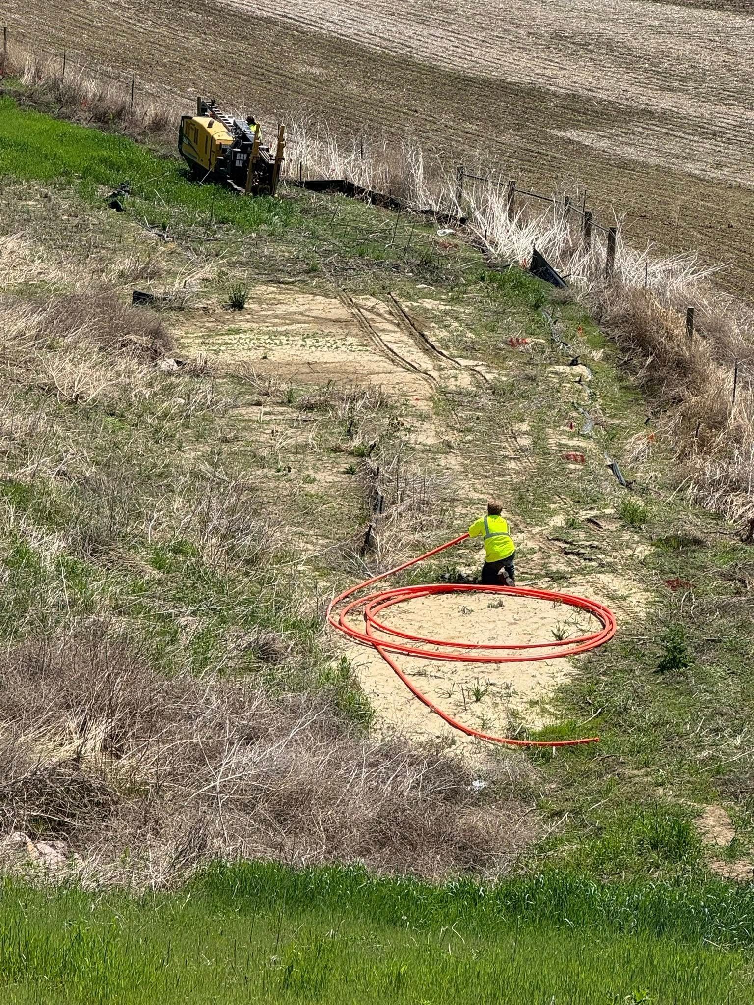 Person in yellow vest working with orange coiled tubing on a hillside, with equipment nearby.