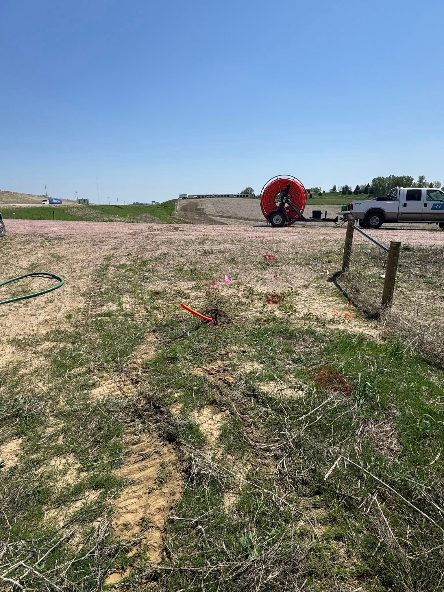 Construction site with red utility line reel, utility truck, and orange flags on a gravel and grass field.