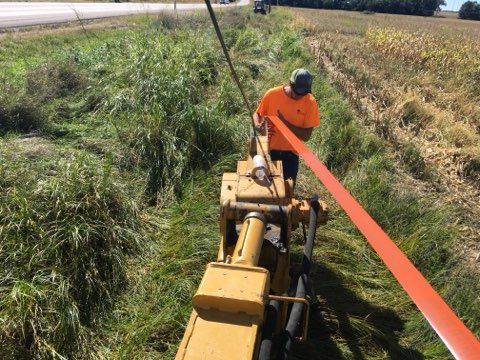 Man operating trenching machine, pulling orange cable through ditch near road and cornfield.