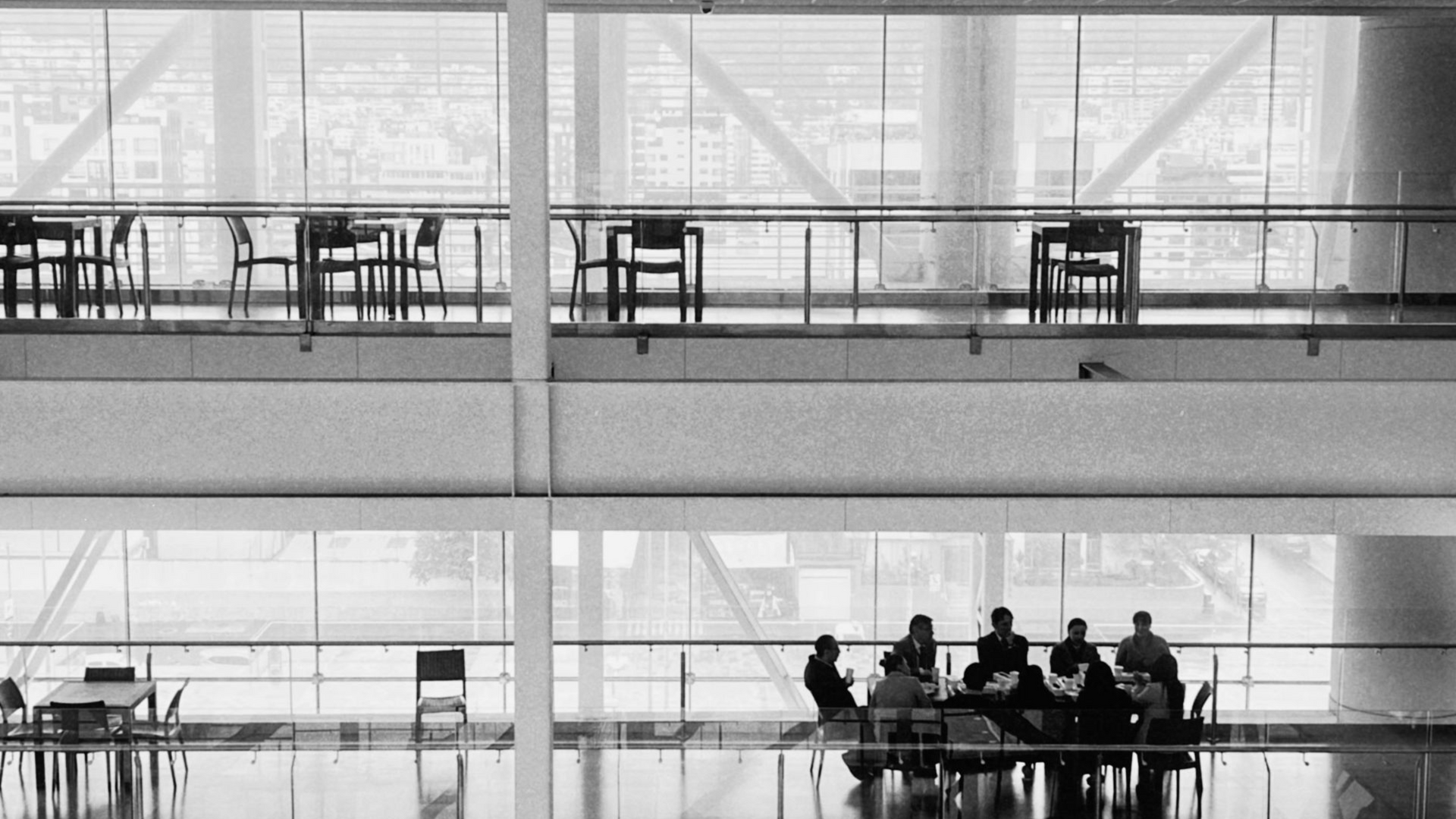 Black and white photo of a modern building interior. People sit around tables on the ground floor, tables and chairs are visible on an upper level.