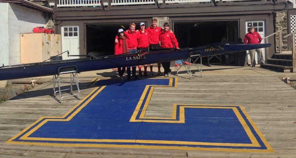 Rowing team in red jackets stands with their boat at a boathouse dock, over a large blue 