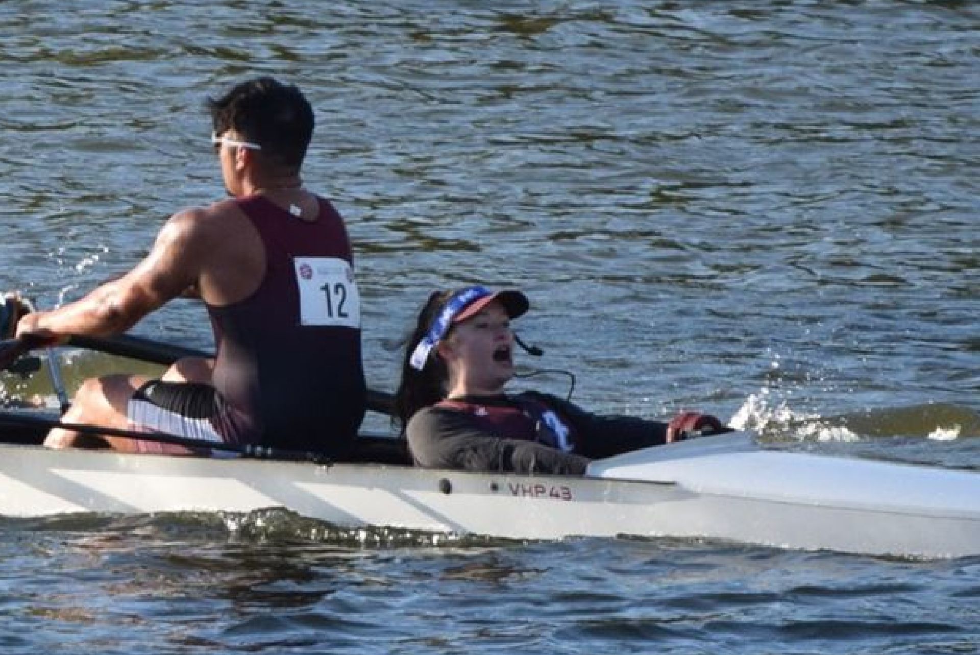 Two rowers in a white boat, paddling on water. The man in the back pulls with effort, the woman in front looks to be yelling.