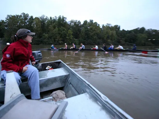 A person in a small motorboat watches a crew team rowing on a river. The sky is overcast, and trees line the bank.