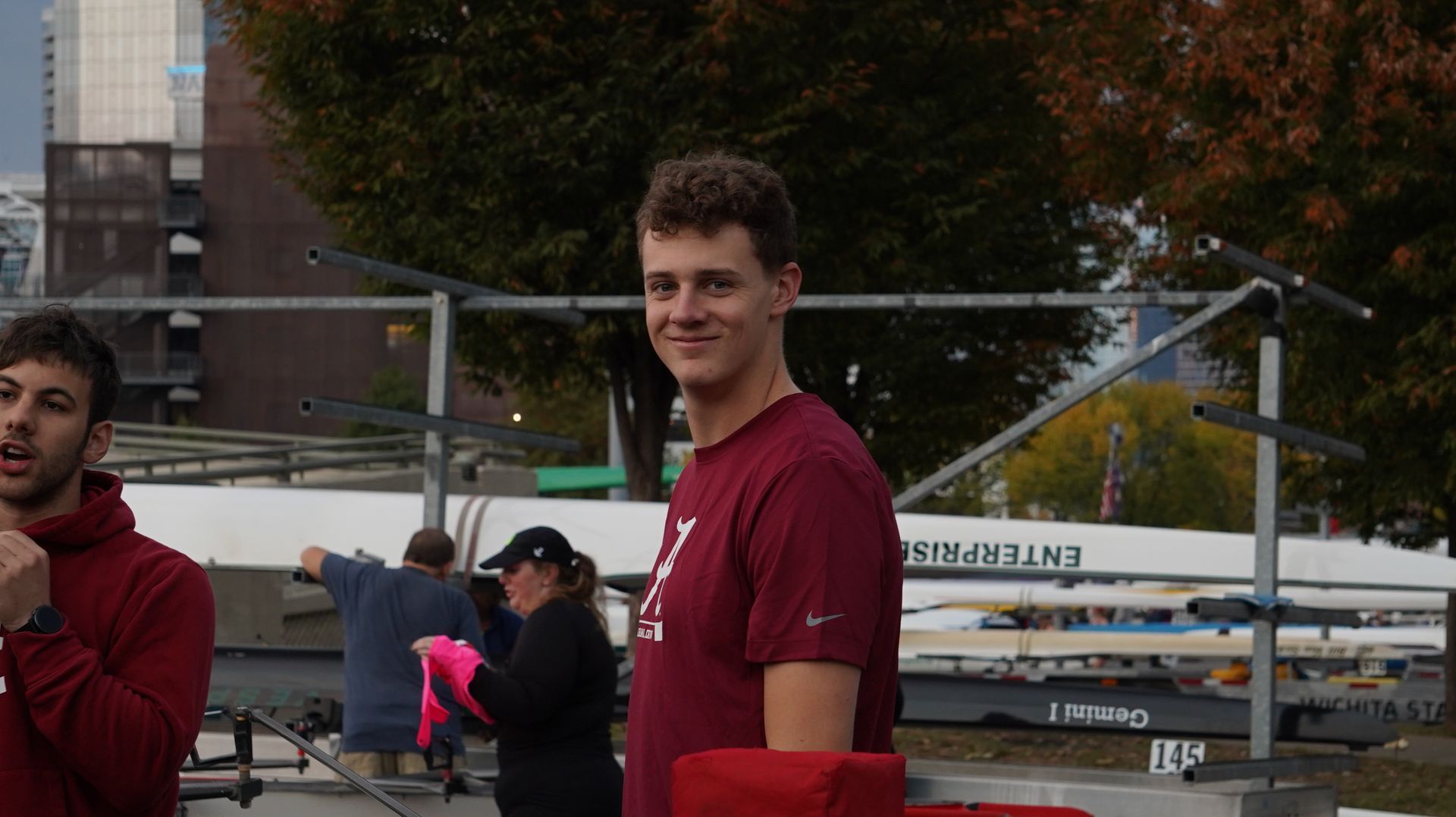 Man in maroon shirt smiles, standing near a rowboat and other people outdoors.