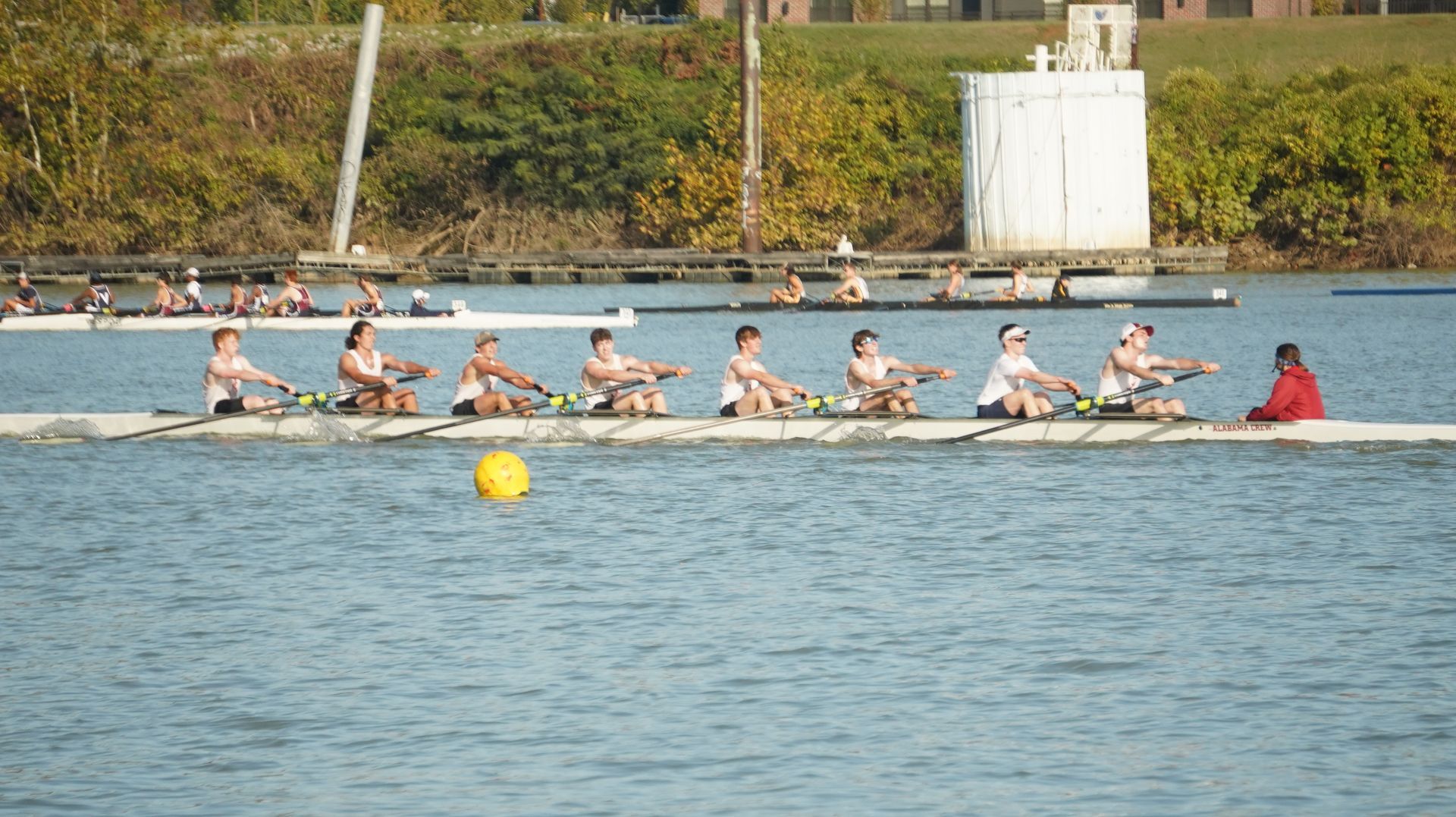 Rowing team on a long boat, with eight rowers in unison, on a river. Coach in red jacket sits in the back.