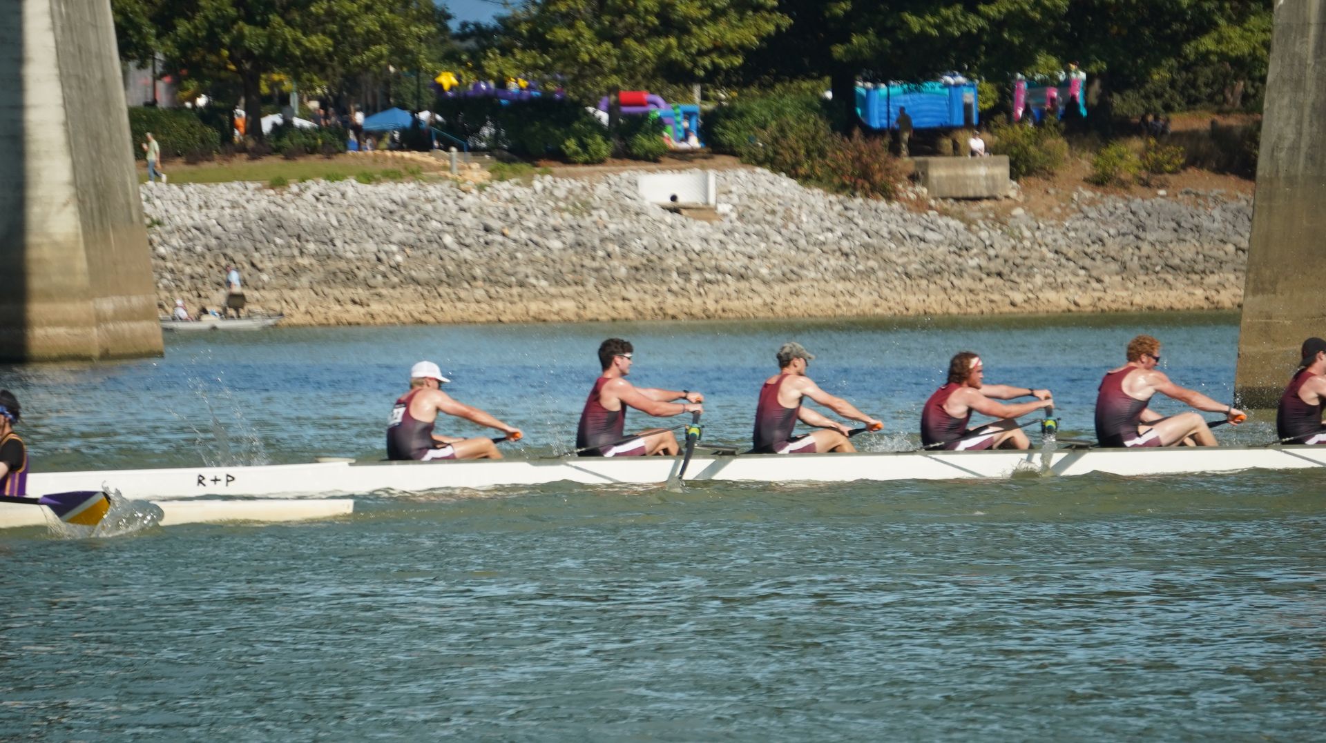 Rowing team in a white boat, rowing on a river. They are wearing dark colors, with a bridge in the background.