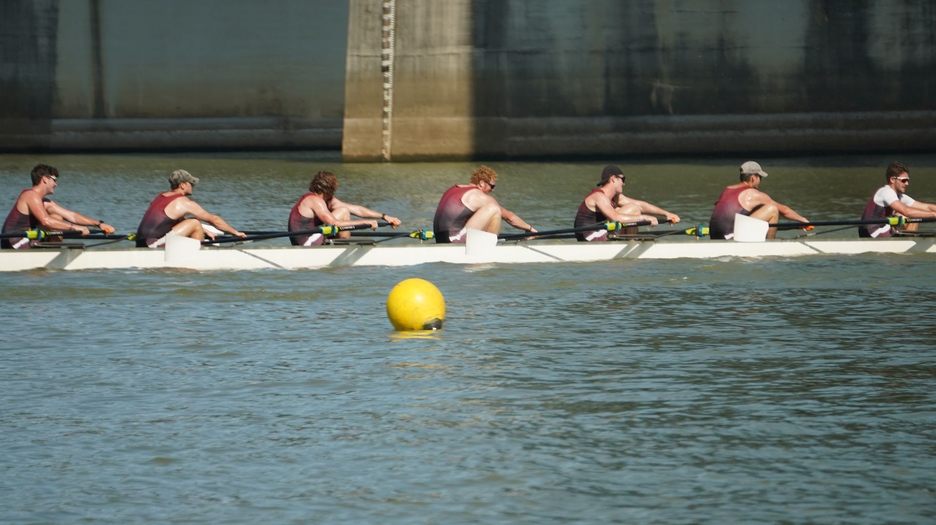 A crew team of eight rowers on a body of water, rowing towards the camera. The rowers are wearing maroon and black uniforms.