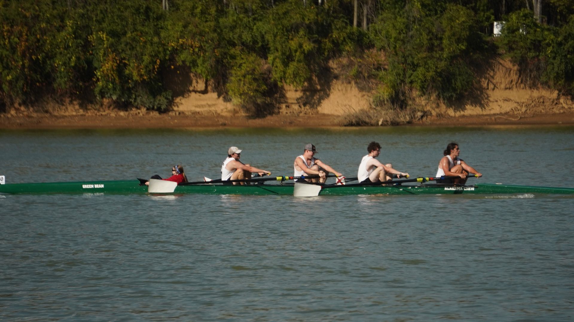 A crew team in a green boat rowing on a river. Trees line the bank.