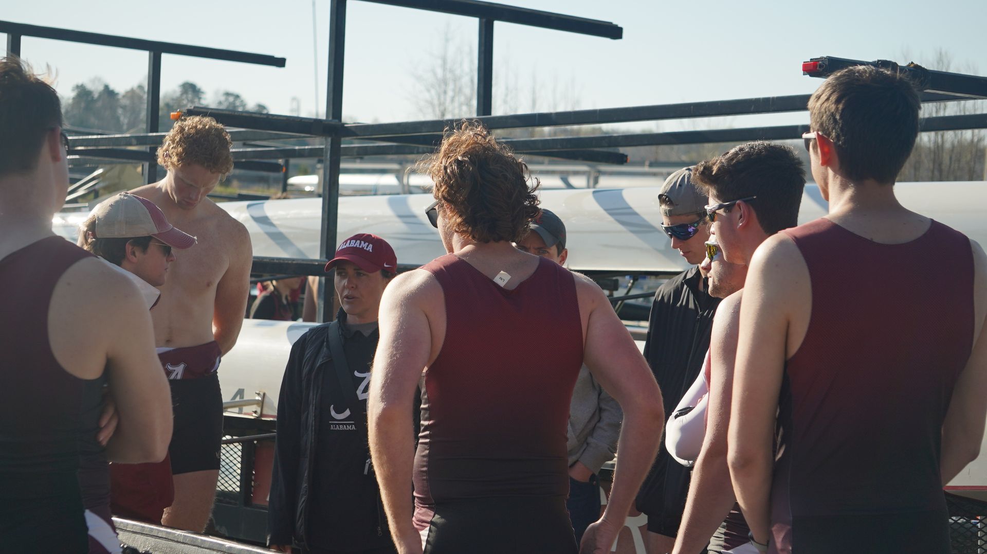 A group of rowers in maroon singlets gather outdoors, likely before or after a race, with the boathouse in the background.