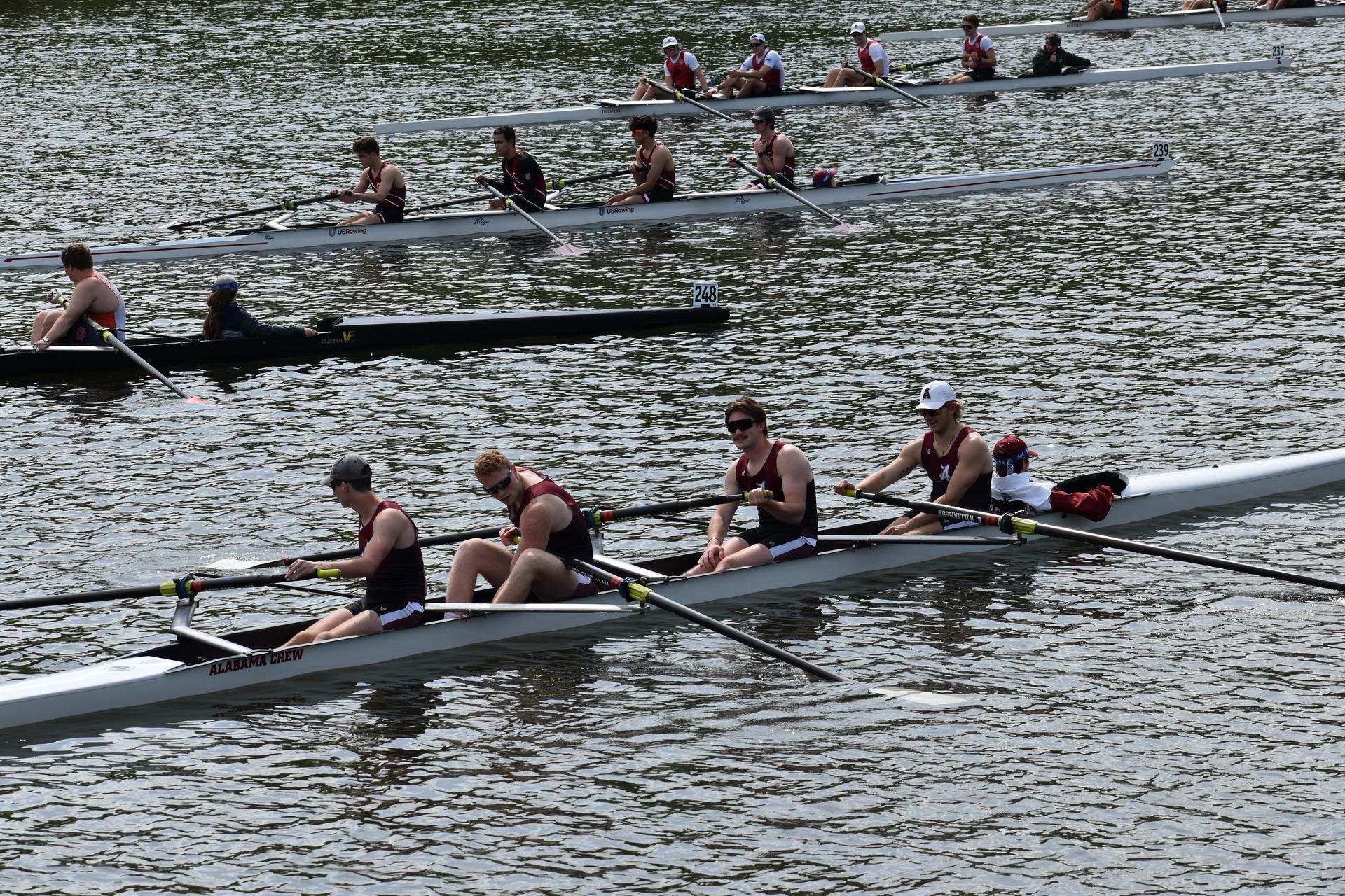 Rowing teams in boats on a body of water, racing. Athletes wear dark uniforms, sunlight reflects on the water.