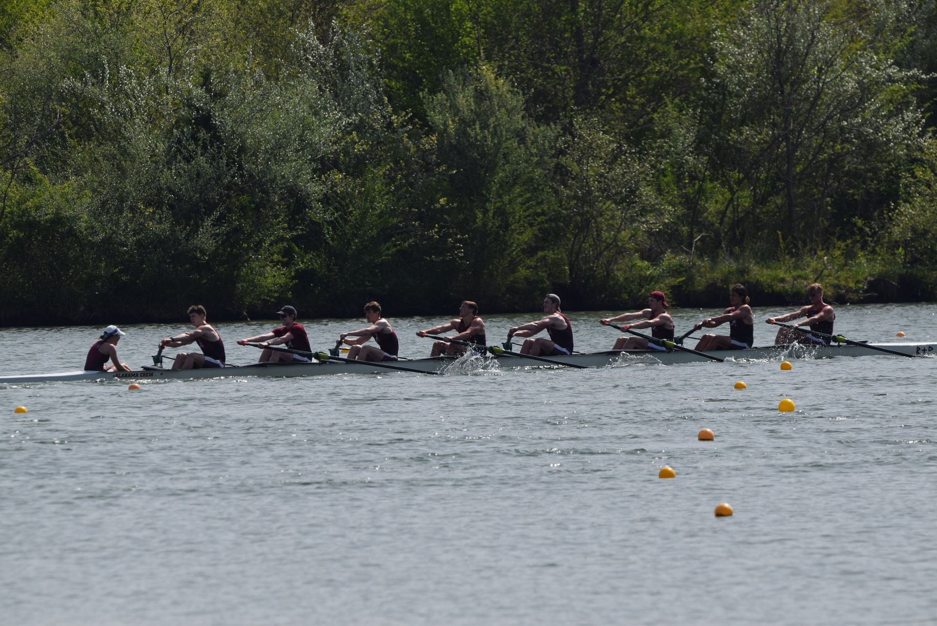 A rowing crew of eight athletes races on a river under a sunny sky. The rowers are in dark uniforms.