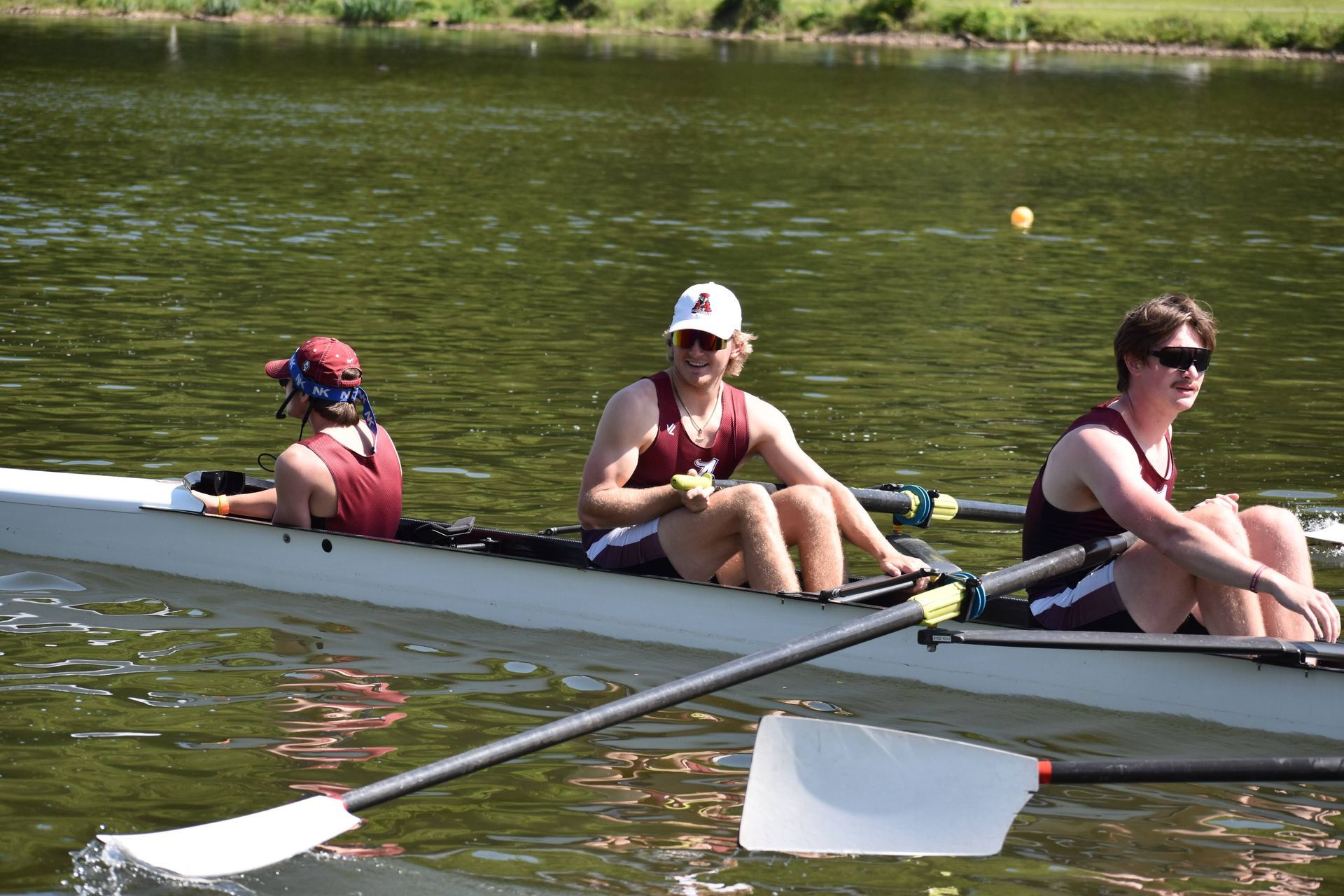 Three rowers in a boat on a lake, wearing maroon and white athletic gear. One wears a white cap, all are focused on rowing.